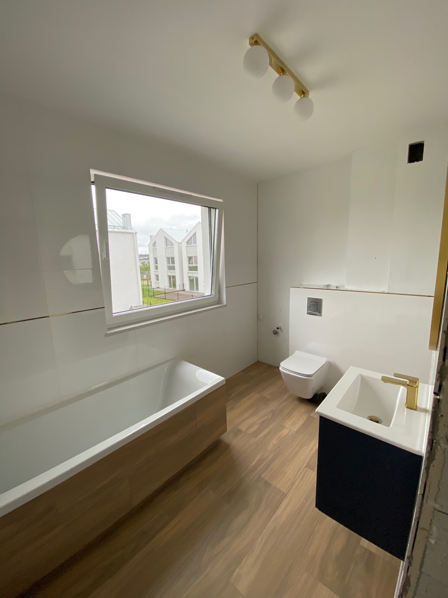 Wnętrze łazienki w trakcie wykańczania: białe płytki na ścianach, drewnopodobna podłoga, wanna obudowana drewnem, umywalka z czarną szafką i złotą armaturą, toaleta, widok z okna na budynki...