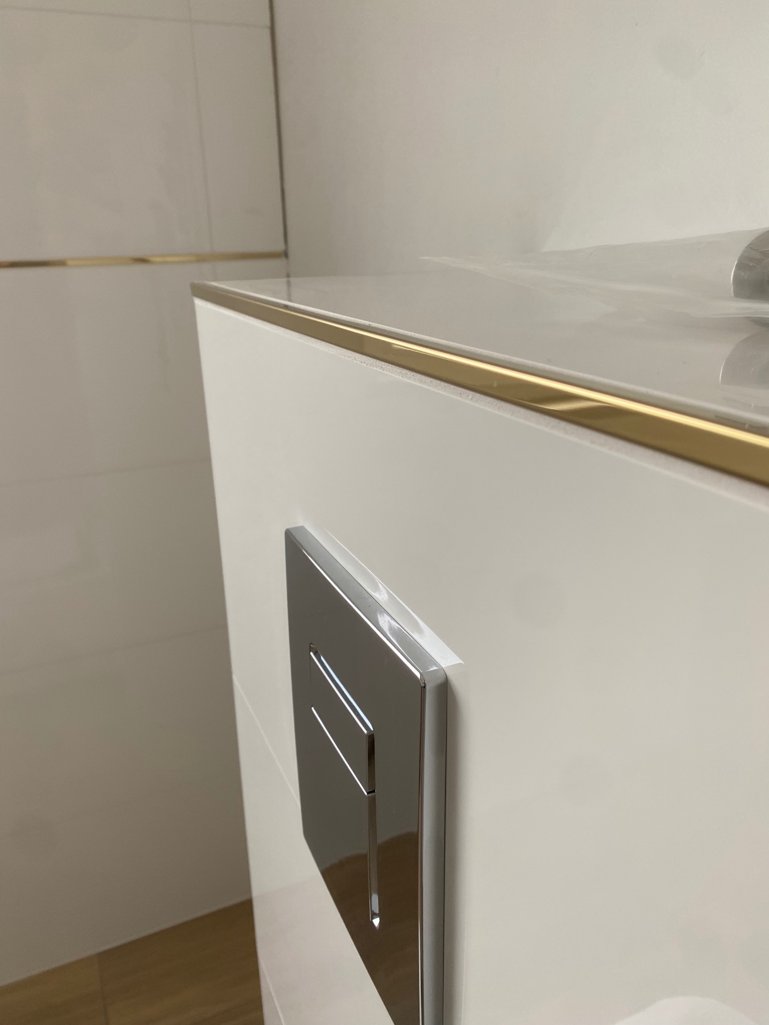 Biała zabudowa stelaża WC z chromowanym przyciskiem spłukującym, widoczna złota listwa dekoracyjna na styku zabudowy i płytek ściennych.