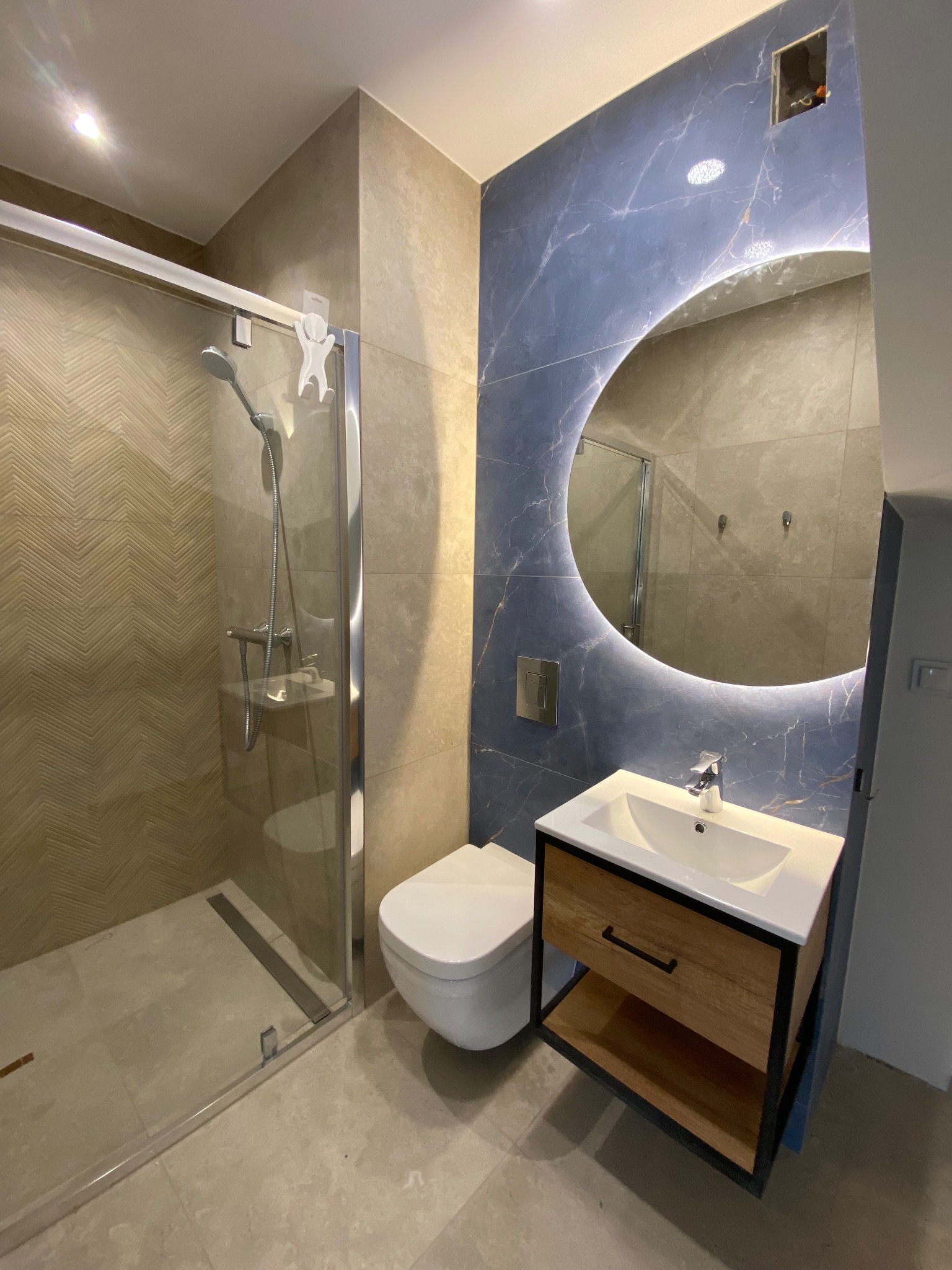 Nowoczesna łazienka z kabiną prysznicową, umywalką z drewnianą szafką na czarnym stelażu, okrągłym lustrem z podświetleniem LED i niebieskimi płytkami imitującymi marmur na ścianie.