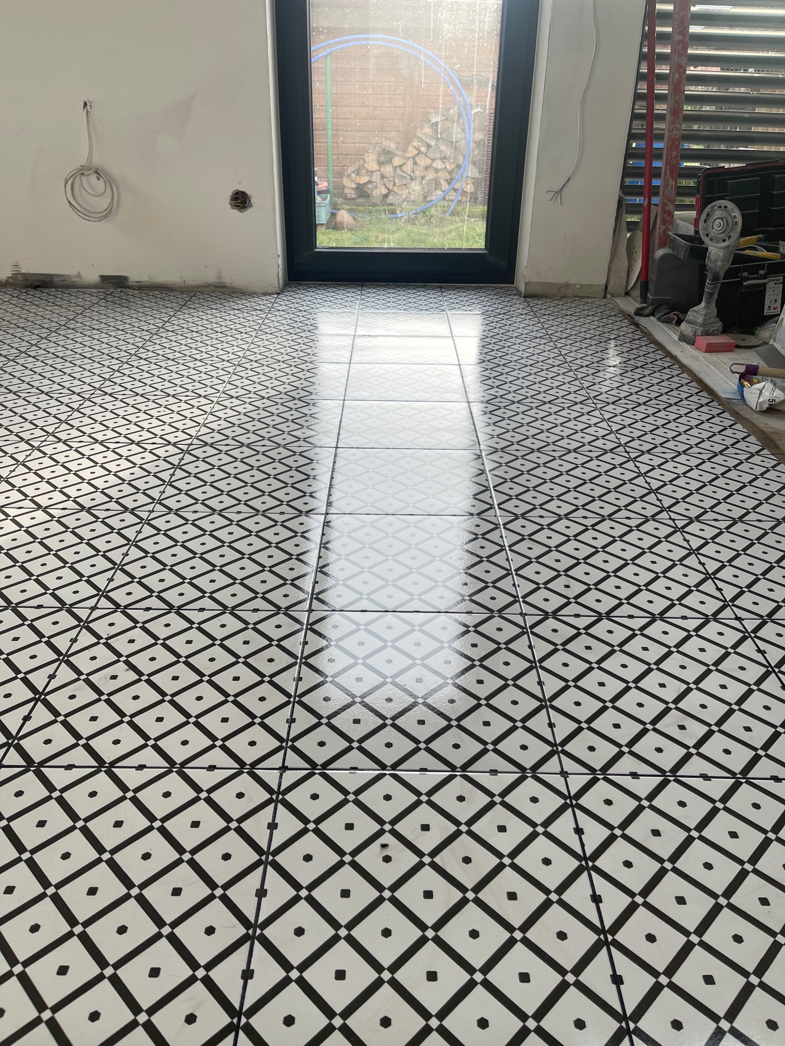 Ułożona podłoga z błyszczących, czarno-białych płytek w geometryczny wzór, widoczne niedokończone ściany i narzędzia glazurnicze w tle.