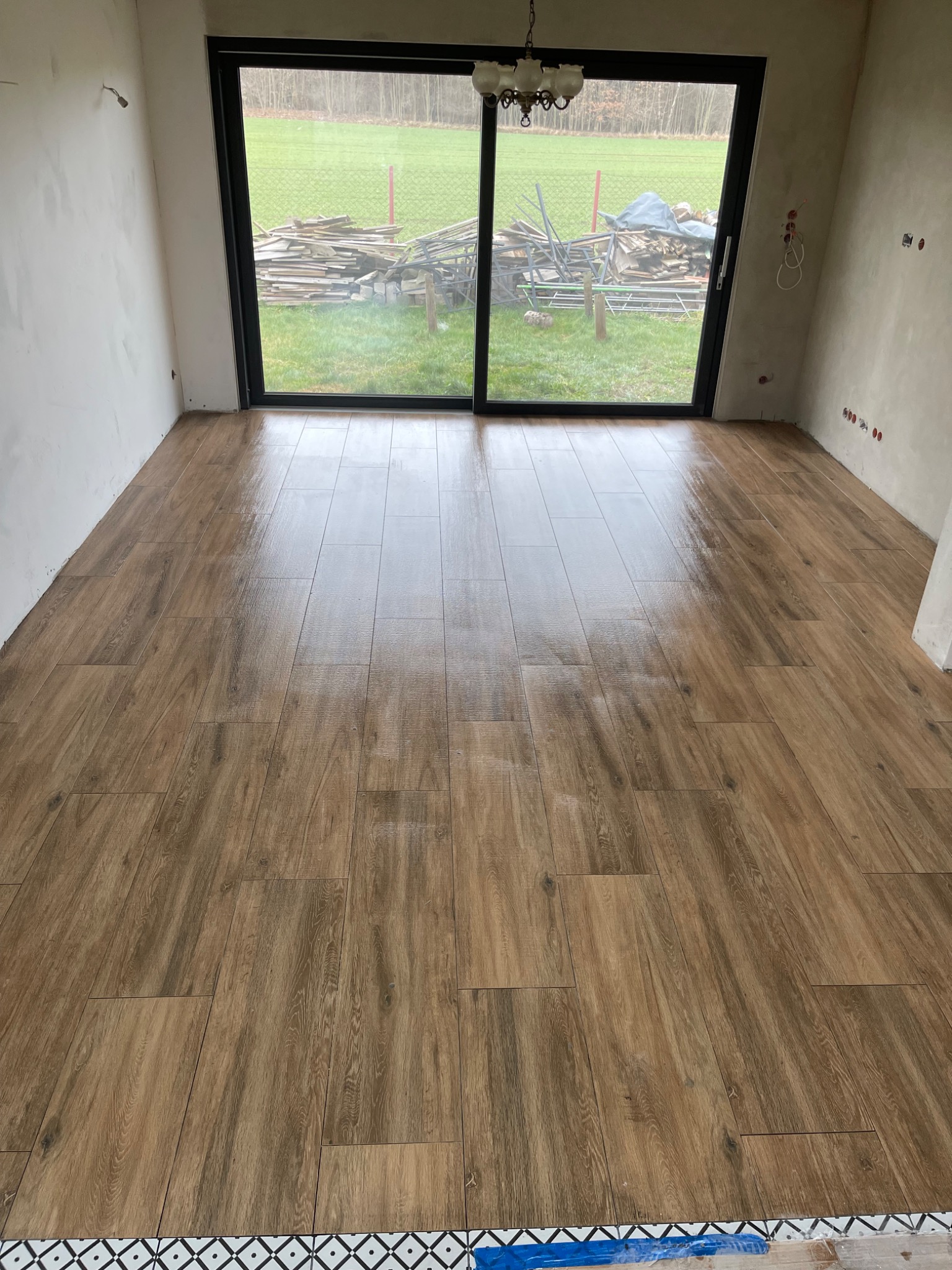 Świeżo ułożona, połyskująca podłoga z płytek imitujących drewno w pustym pomieszczeniu z dużym oknem balkonowym i widokiem na podwórze z ułożonymi deskami; dolna krawędź wykończona dekoracyjną...