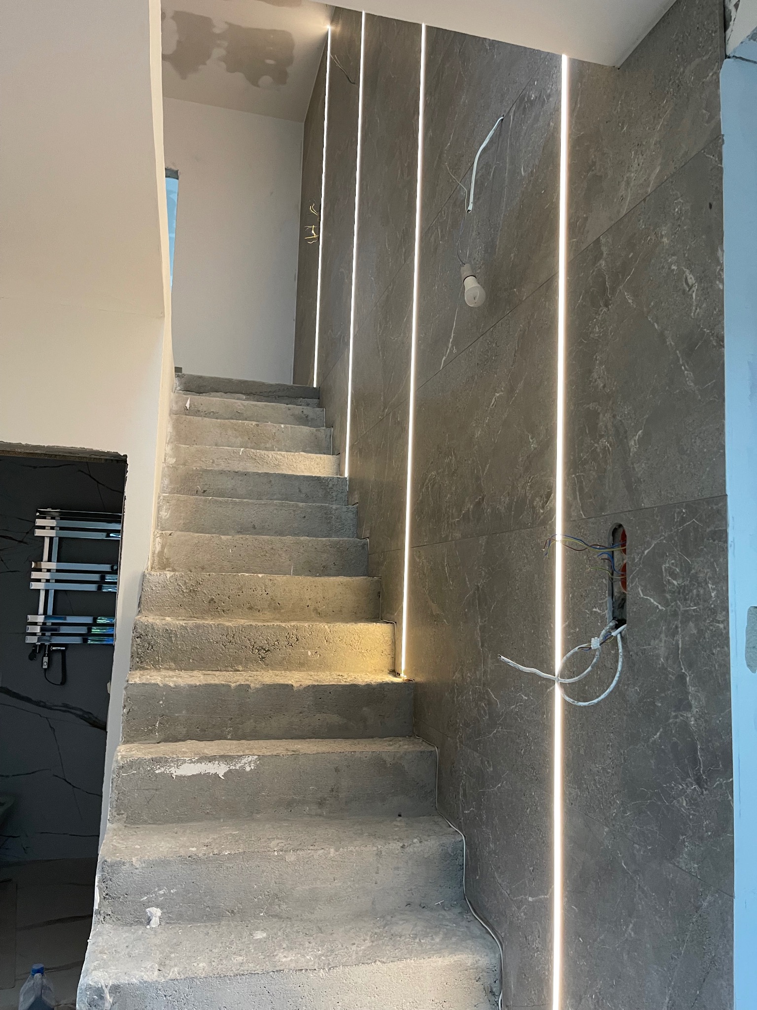 Surowe betonowe schody w trakcie wykańczania, z podświetlanymi pionowymi panelami ściennymi imitującymi marmur i wystającymi kablami elektrycznymi.