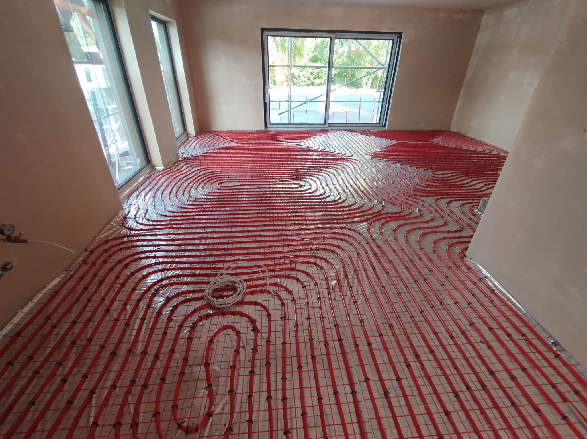 Czerwone rury ogrzewania podłogowego ułożone spiralnie na podkładzie w surowym pomieszczeniu z oknami, widoczne zwoje rury.