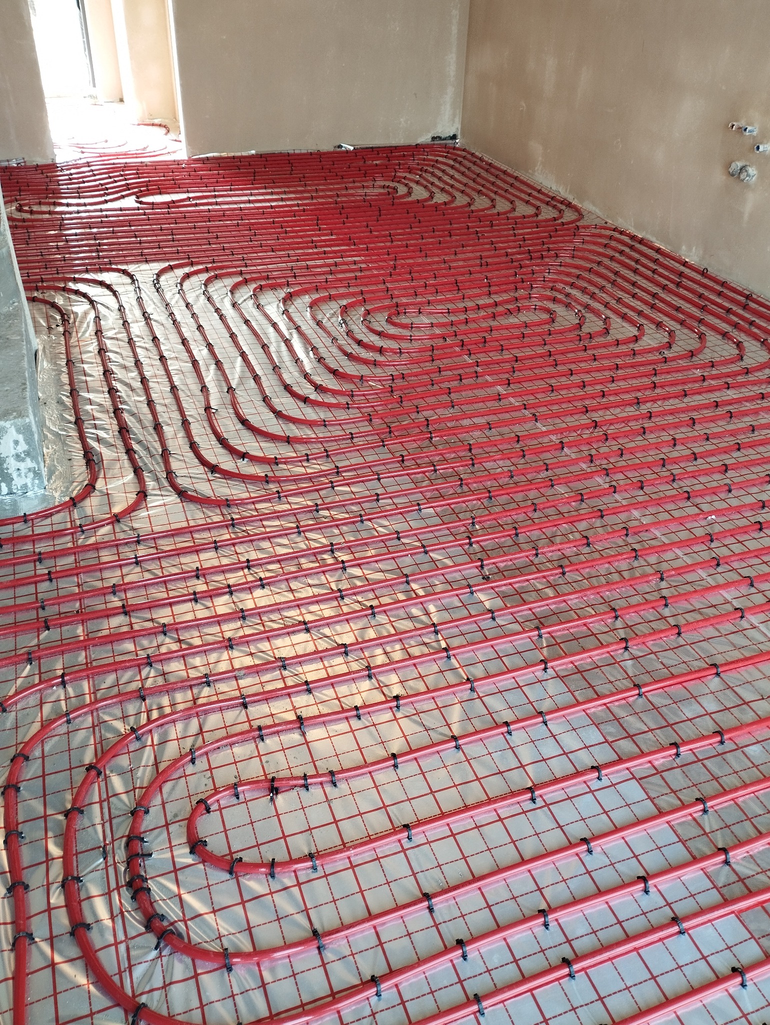 Czerwone rury ogrzewania podłogowego ułożone w pętle na folii z siatką, przymocowane czarnymi uchwytami, widoczne szare ściany i fragment podłogi w surowym stanie.