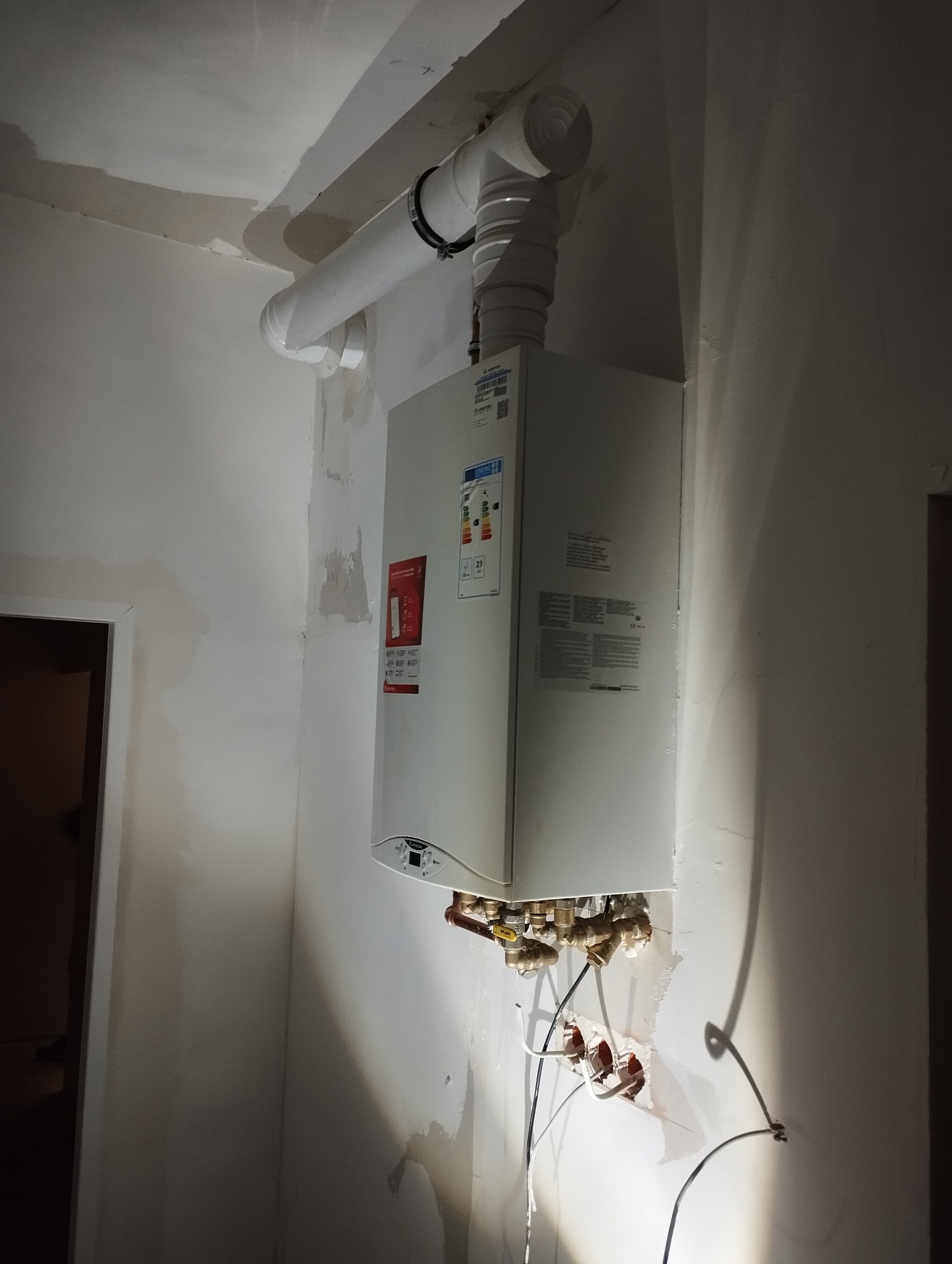 Nowy piec gazowy marki Ariston zamontowany na ścianie w surowym stanie, widoczne rury odprowadzające spaliny i instalacja hydrauliczna z licznymi zaworami, ściany wymagają wykończenia.