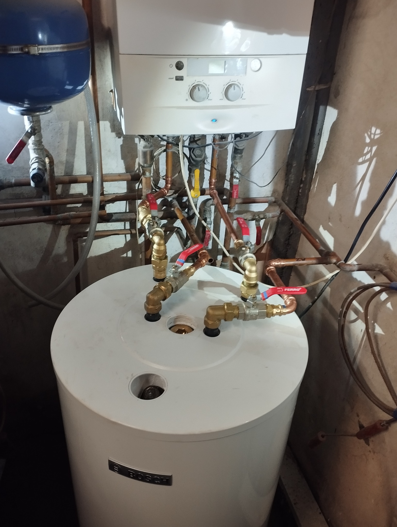 Biały zasobnik ciepłej wody użytkowej Bosch z podłączonymi rurami miedzianymi i zaworami z czerwonymi rączkami, zamontowany pod wiszącym piecem gazowym i niebieskim zbiornikiem wyrównawczym...