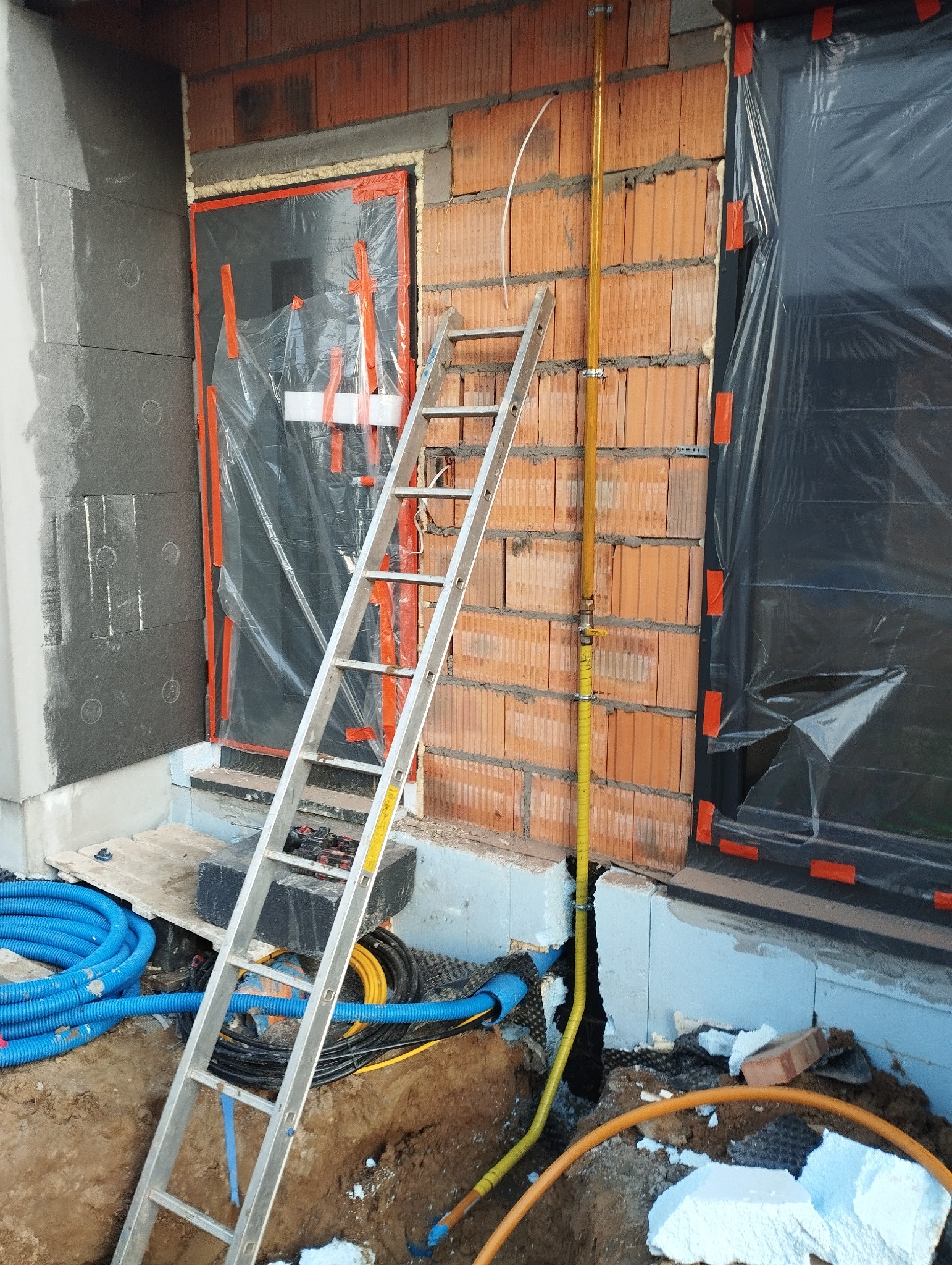 Żółta rura gazowa przymocowana do ściany z cegły na zewnątrz budynku, widoczna drabina aluminiowa, niebieskie rury karbowane i fragment wykopu.