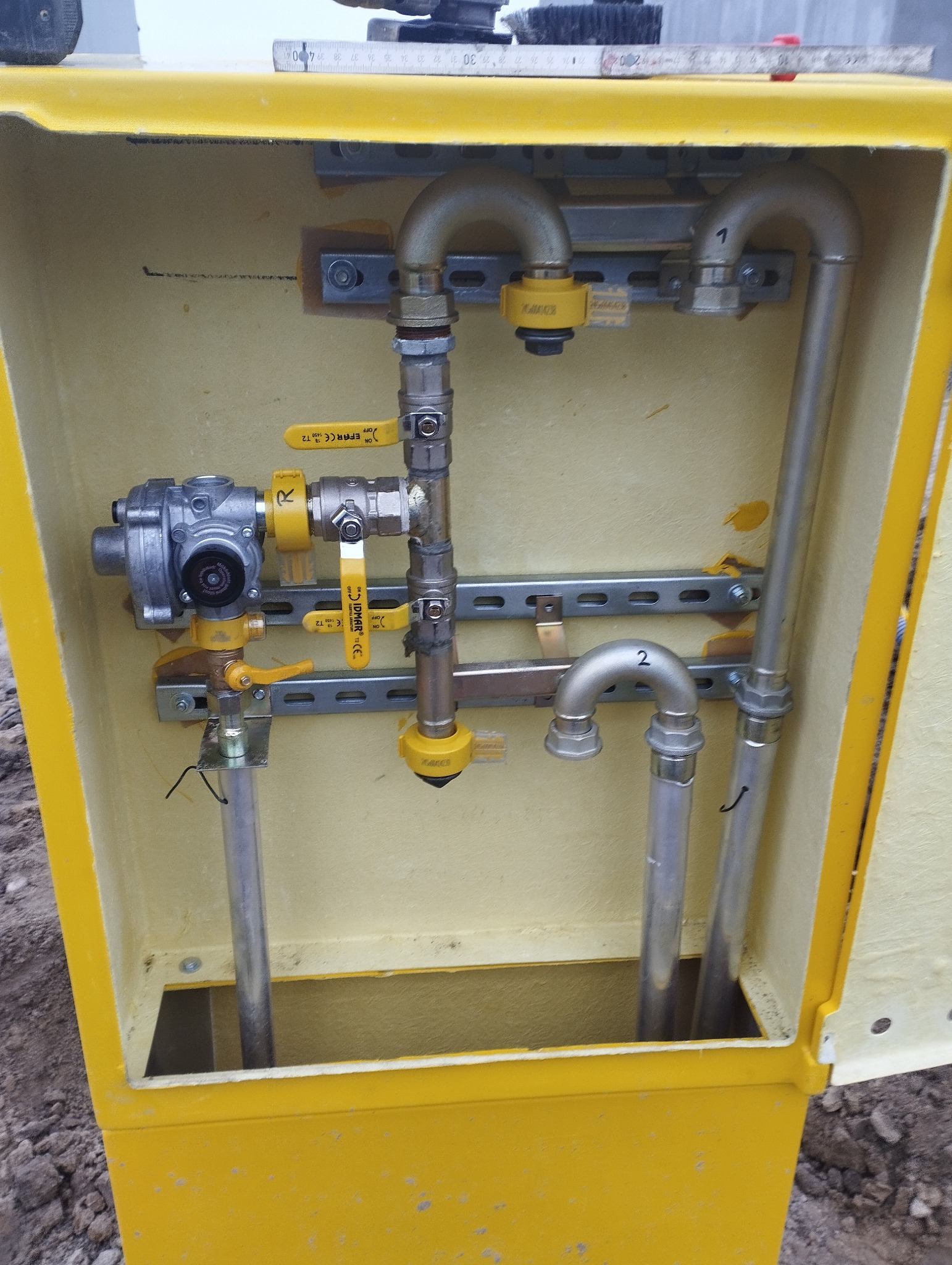 Żółta skrzynka z zamontowaną instalacją gazową, widoczne zawory z żółtymi uchwytami i rury z oznaczeniami 1 i 2, szyny montażowe.