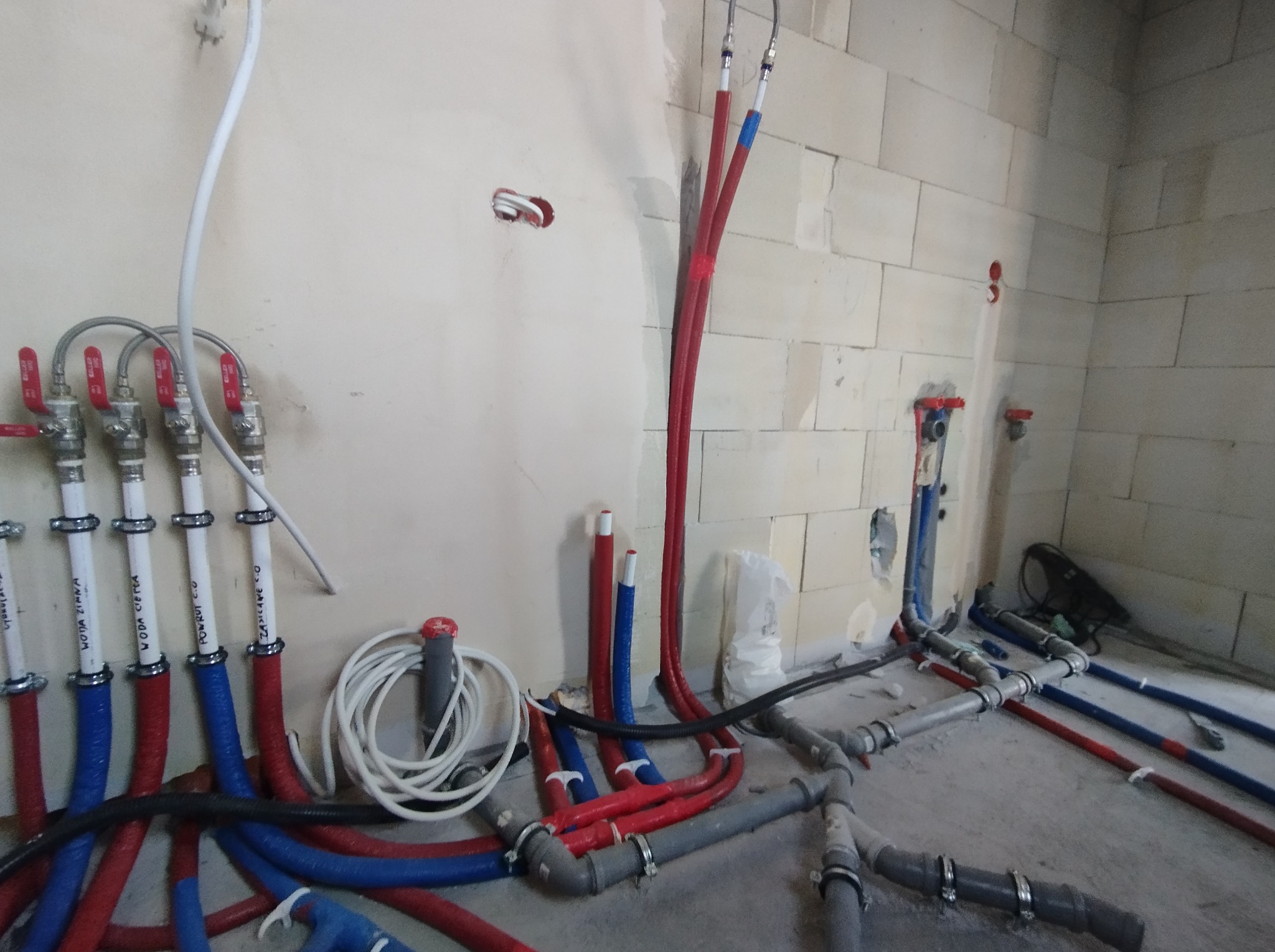 Instalacja hydrauliczna w trakcie montażu: rury w izolacji czerwonej i niebieskiej, szare rury kanalizacyjne, zawory oraz przewody elektryczne na tle surowej ściany z betonu komórkowego.