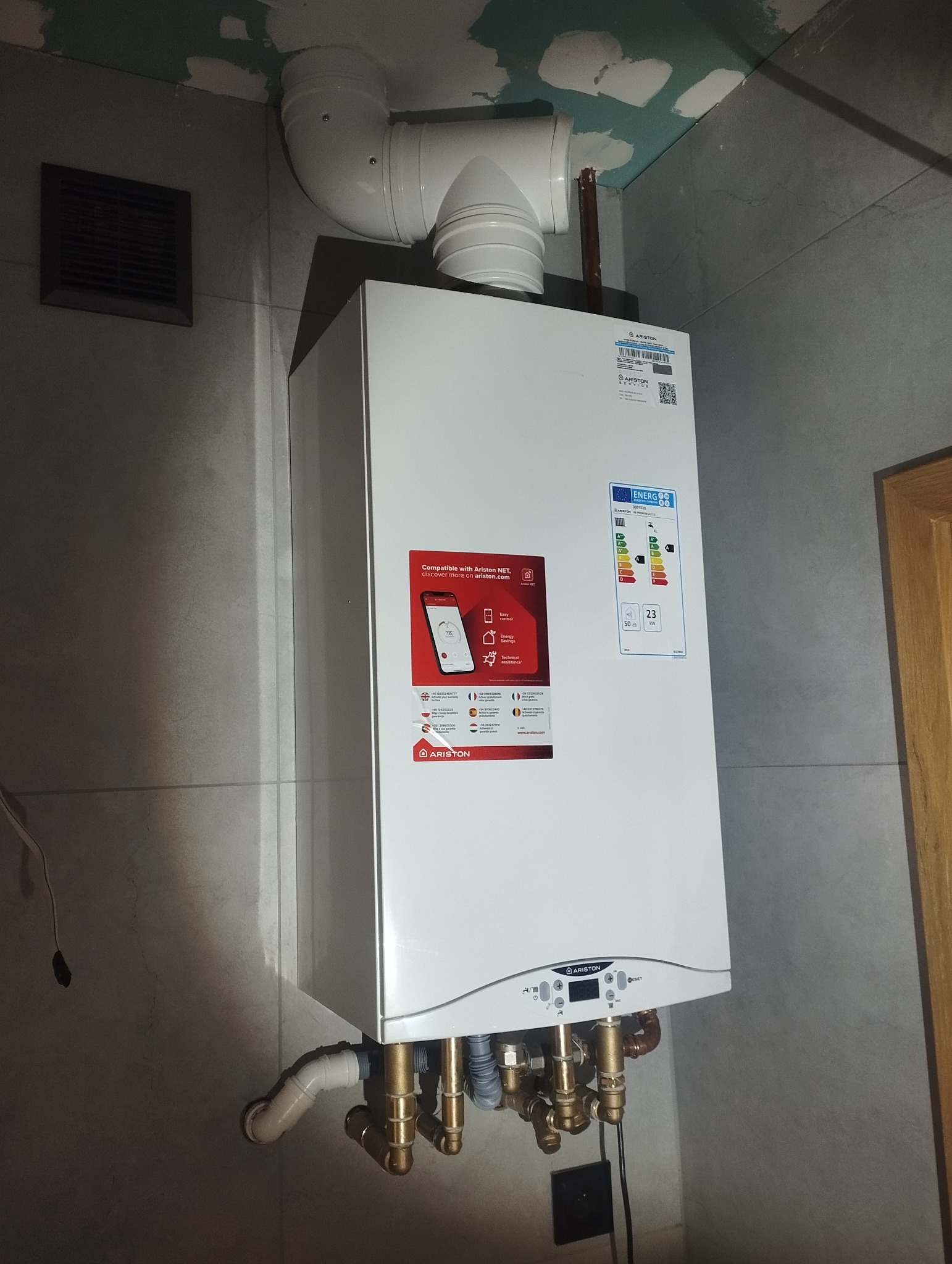 Nowo zamontowany, biały piec gazowy Ariston z widocznymi rurami odprowadzającymi spaliny i przyłączami wodnymi, zamontowany na szarej ścianie w trakcie prac wykończeniowych.