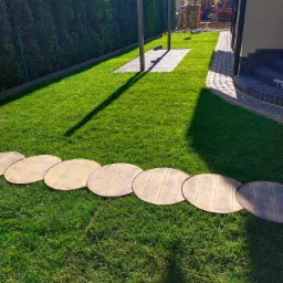 Świeżo skoszony trawnik z okrągłymi płytami chodnikowymi z imitacją drewna, prowadzącymi przez środek, w tle żywopłot z tui i huśtawka.