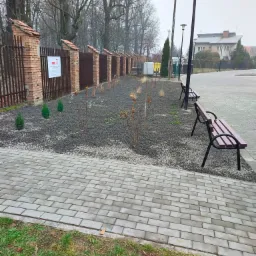 Nowo założony rabat z krzewami i drobnymi iglakami wysypany grysem, obok chodnik z kostki, ławki parkowe i oświetlenie uliczne, w tle budynki.