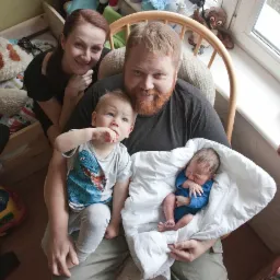 Rodzinne zdjęcie: ojciec z rudą brodą siedzi w fotelu z noworodkiem w białym kocyku i starszym synkiem na kolanach, obok stoi uśmiechnięta mama, a w tle widać pokój dziecięcy z zabawkami.