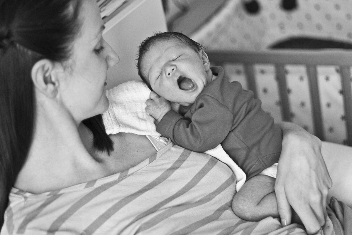Czarno-białe zdjęcie noworodka ziewającego na ramieniu matki, w tle dziecięce łóżeczko.