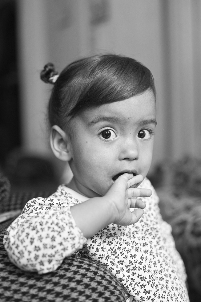Czarno-białe portretowe ujęcie dziecka z palcami w ustach, ubrane w bluzkę w drobne kwiatki, siedzące na tkaninie w pepitkę.