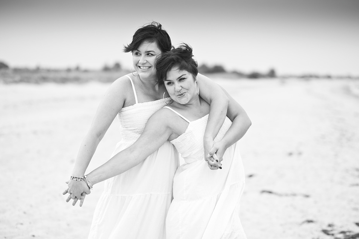 Dwie uśmiechnięte kobiety w białych sukienkach, obejmujące się na plaży w czarno-białej fotografii, z widocznymi kolczykami kołami i bransoletką na nadgarstku jednej z nich.