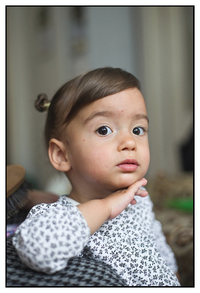 Portret uroczej dziewczynki z brązowymi włosami upiętymi w mały kok, ubrana w bluzkę w drobne kwiatki, delikatnie dotyka dłonią podbródka.