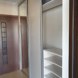 Wnętrze szafy wnękowej z przesuwnymi drzwiami, widoczne białe półki i drążek na ubrania, obok brązowe drzwi wejściowe.