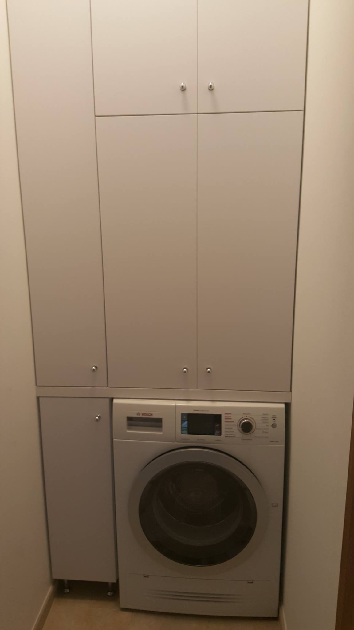 Biała zabudowa meblowa z szafkami nad pralką Bosch w łazience.