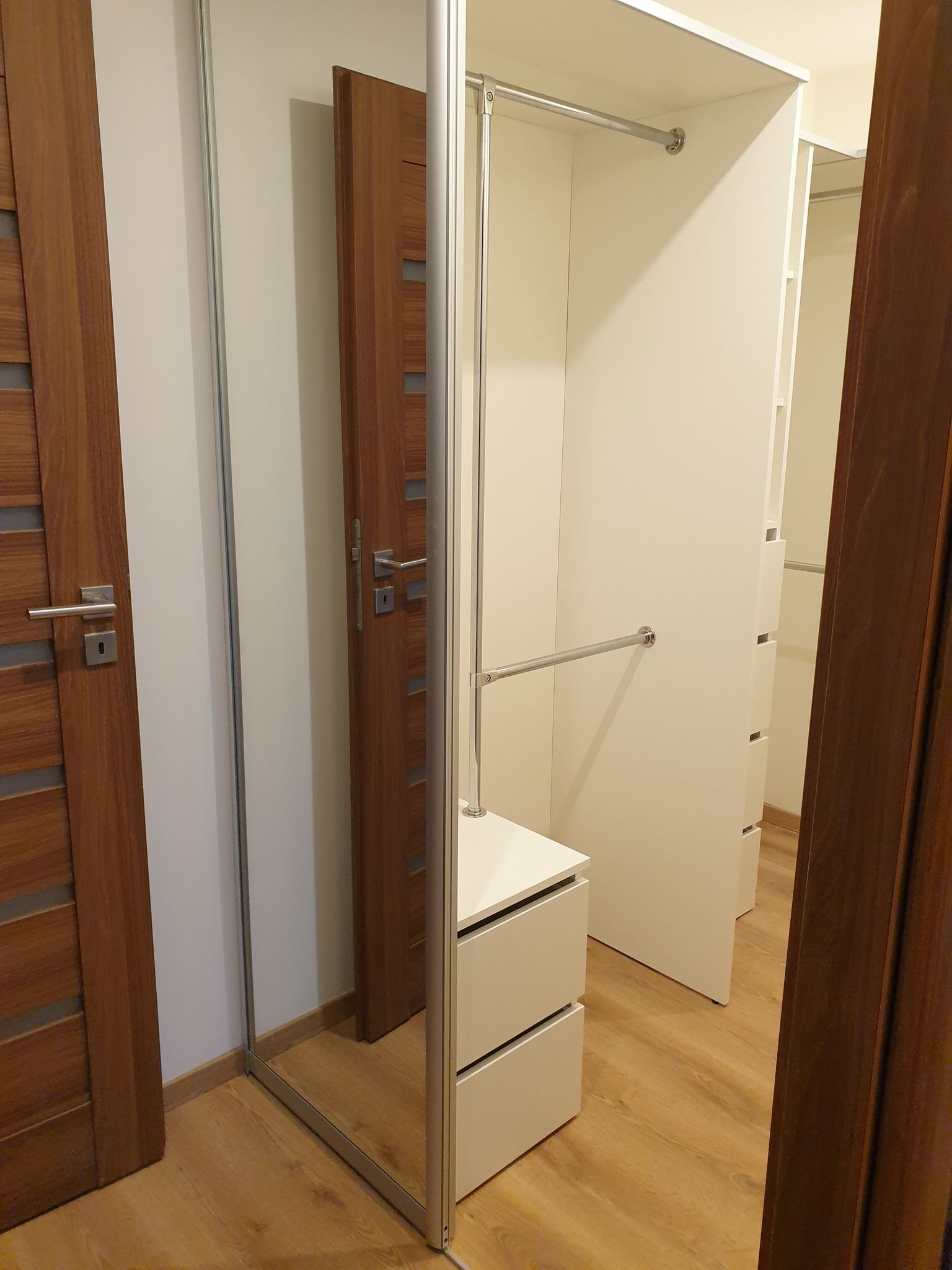 Wnętrze szafy z lustrzanymi drzwiami przesuwnymi, widoczne półki, szuflady i drążki na ubrania; obok drewniane drzwi z poziomymi wstawkami.