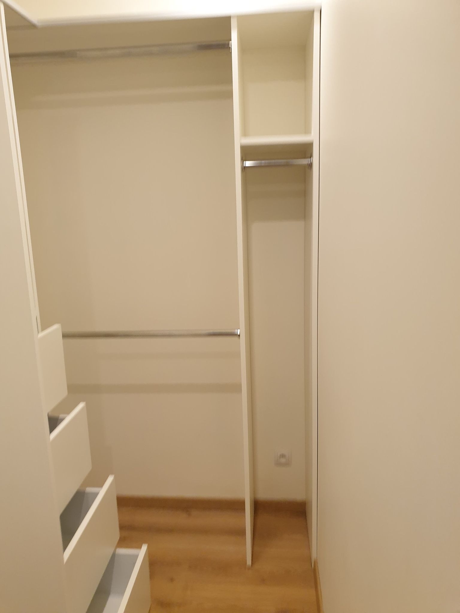 Wnętrze garderoby z białymi frontami szuflad i drążkami na ubrania, widoczne trzy wysunięte szuflady i drewniana podłoga