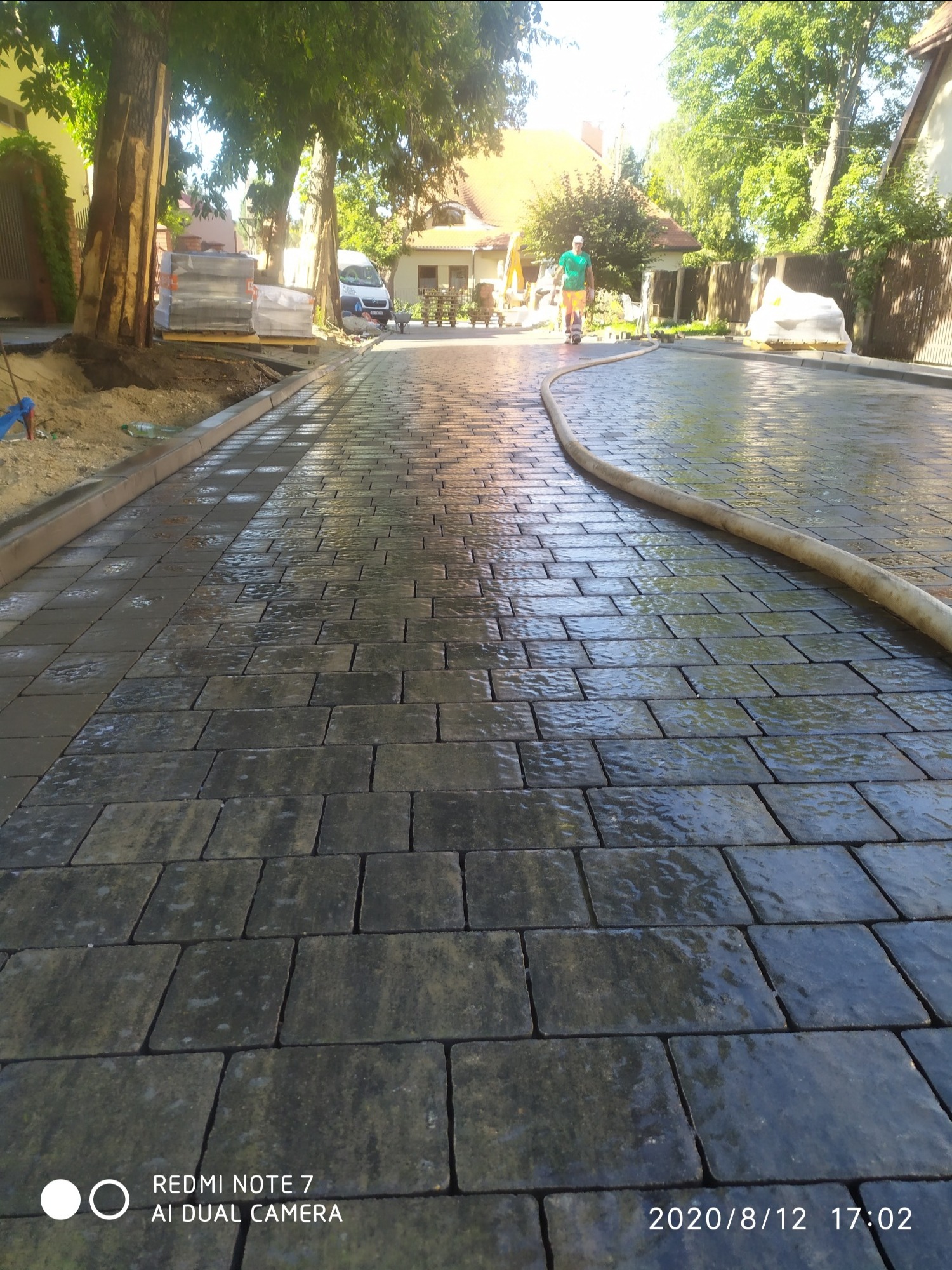 Świeżo ułożona, mokra kostka brukowa w odcieniach szarości, widok z perspektywy chodnika, z widocznym w tle mężczyzną na hulajnodze i stertą kostki czekającą na ułożenie.