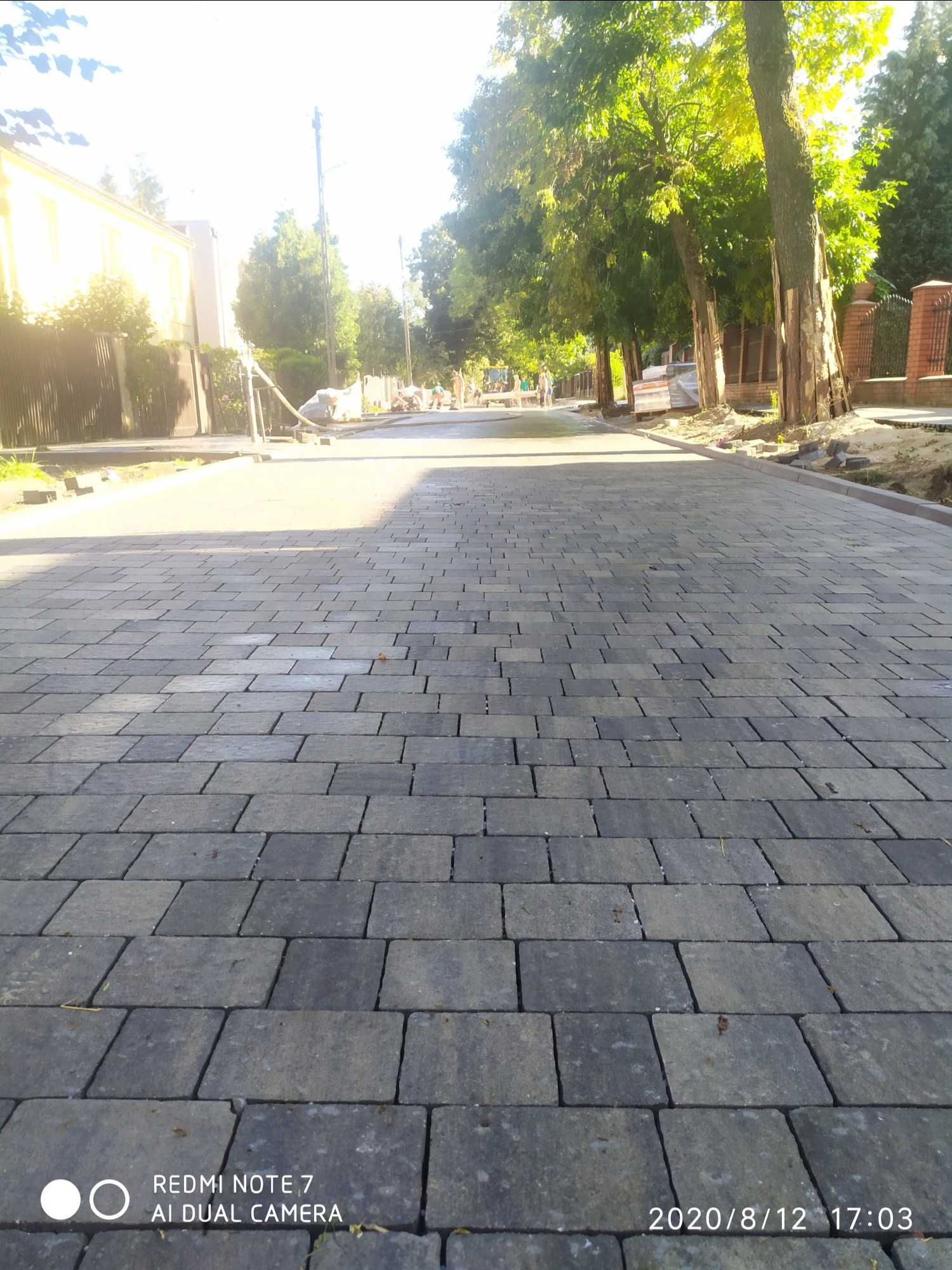 Nowo ułożona, szara kostka brukowa na ulicy w słoneczny dzień, z widocznymi pracownikami w oddali i drzewami po bokach.