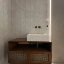 Nowoczesna łazienka z umywalką z lastryko na drewnianej szafce z rattanowymi frontami. Ściana wyłożona płytkami w jodełkę i pionowe oświetlenie LED.