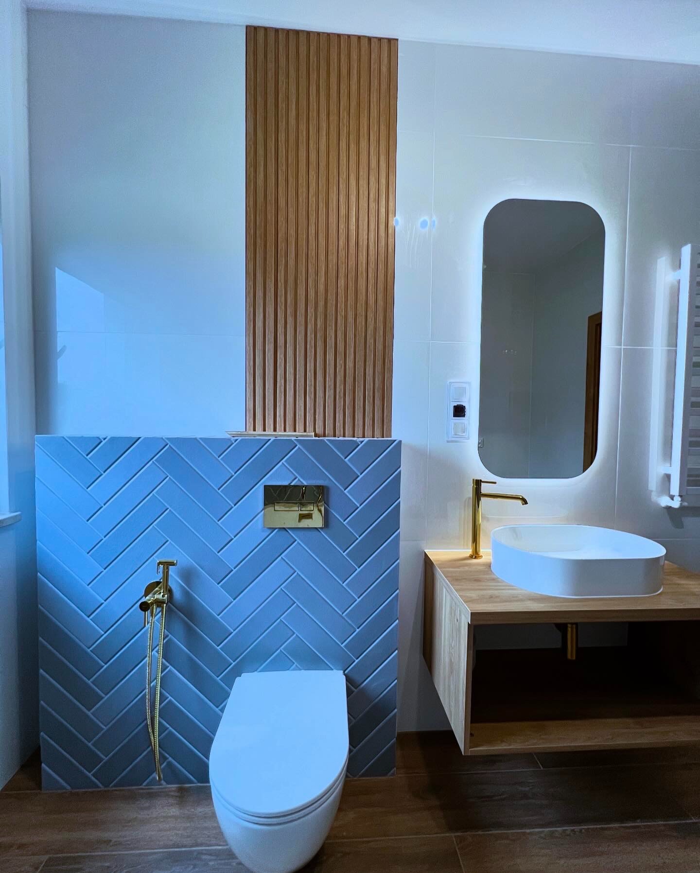 Nowoczesna łazienka z niebieskimi płytkami ułożonymi w jodełkę, drewnianymi akcentami i podświetlanym lustrem. Armatura w kolorze złotym.