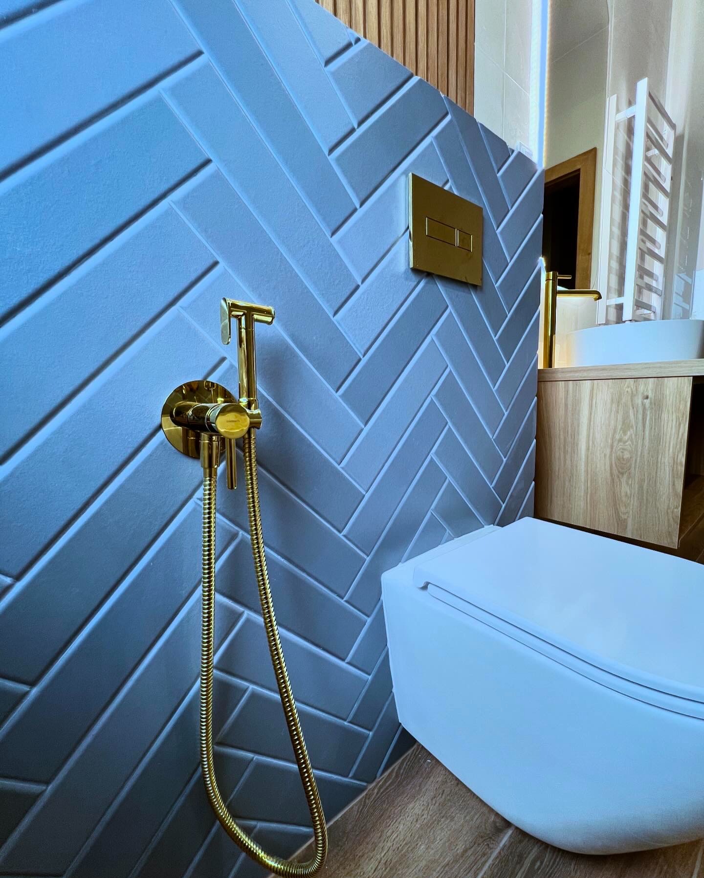 Stylowa łazienka z szaro-niebieską ścianą z płytek ułożonych w jodełkę, złotą armaturą i białą miską WC. Widoczny fragment drewnianej szafki pod umywalką i grzejnika.