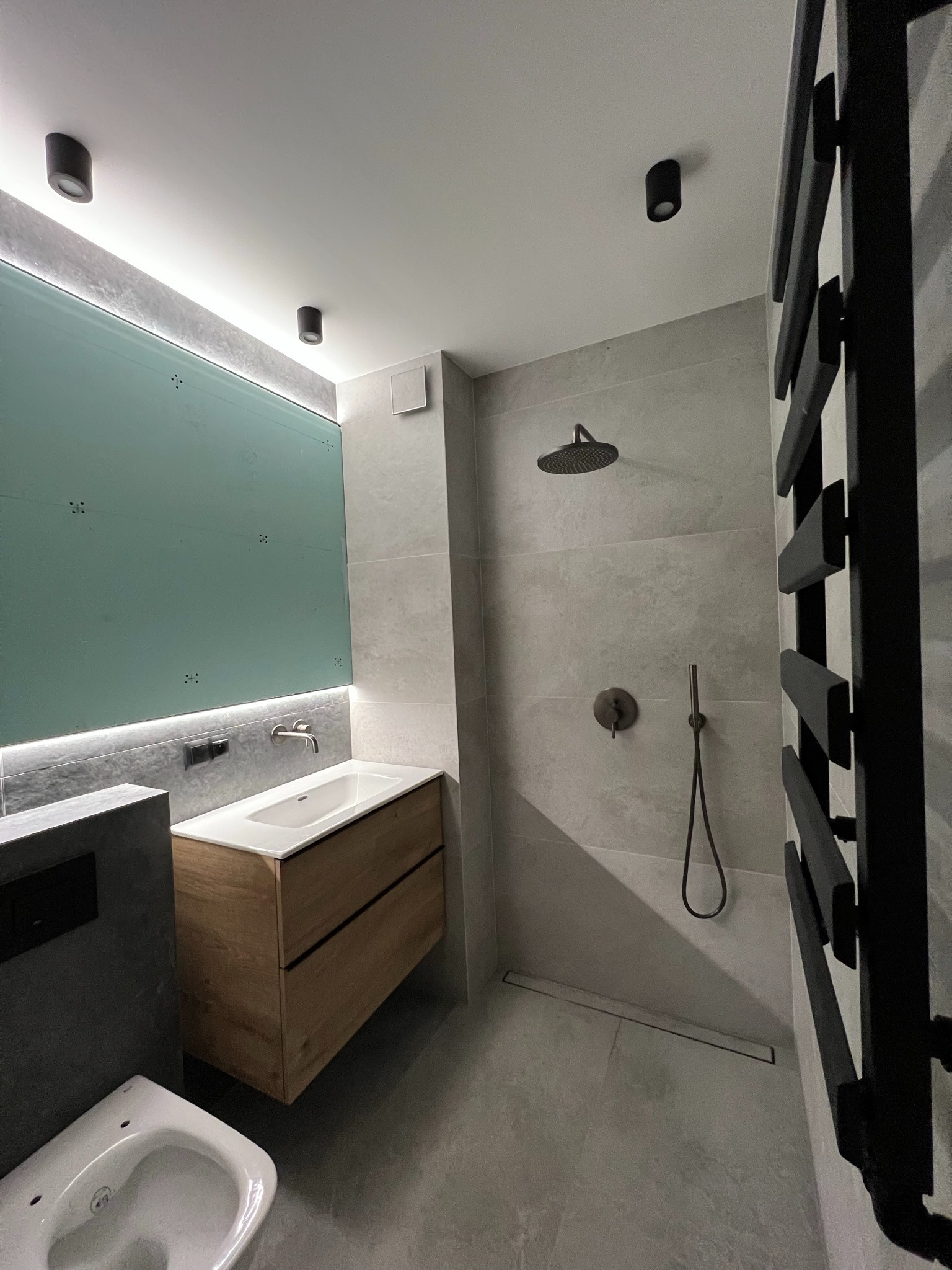 Nowoczesna łazienka z kabiną prysznicową typu walk-in, drewnianą szafką pod umywalką i podświetlanym lustrem. Minimalistyczny design z szarymi płytkami i czarnymi akcentami.