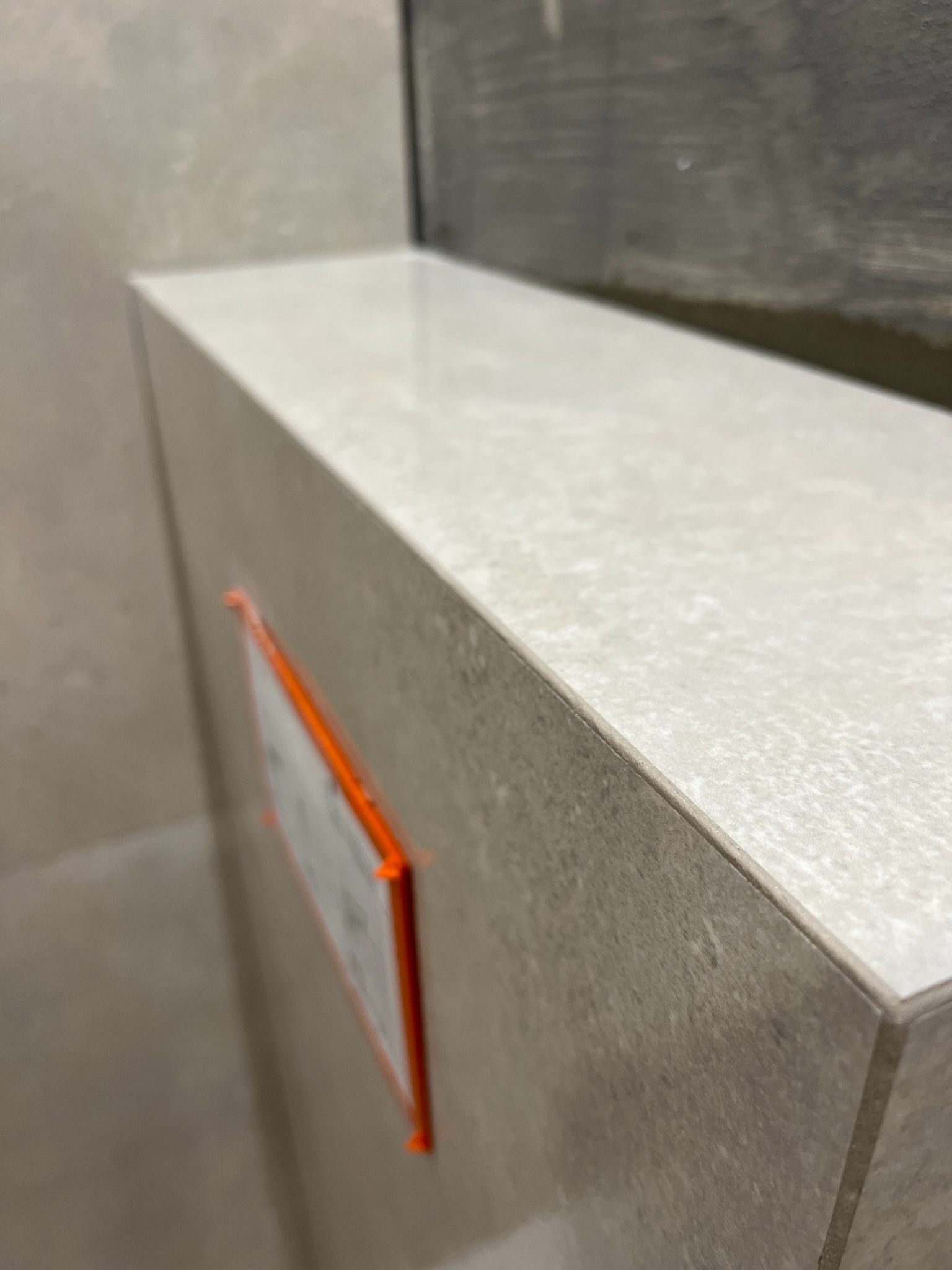 Detal prezentuje wykończoną, jasną, ceramiczną obudowę WC w łazience, z widocznymi pomarańczowymi klinami dystansowymi. Minimalistyczny design, neutralna kolorystyka płytek.