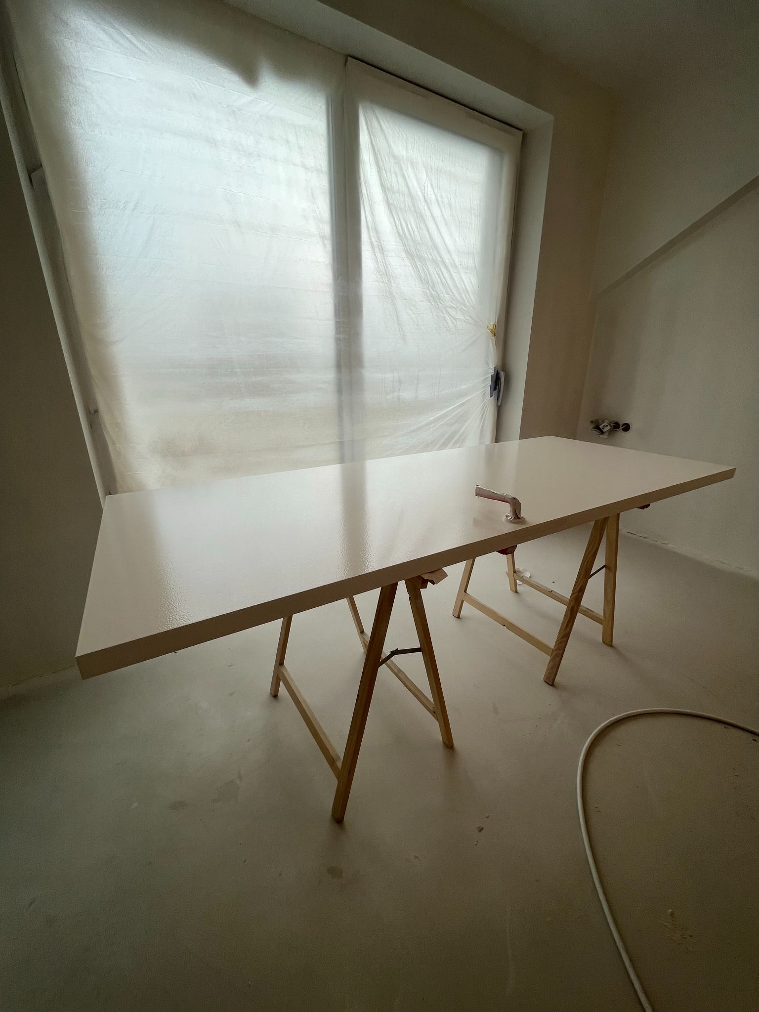 Biały blat na kozłach w pokoju po malowaniu, okno zakryte folią. Świeżo pomalowana powierzchnia odbija światło, w tle ściany w jasnym odcieniu beżu.