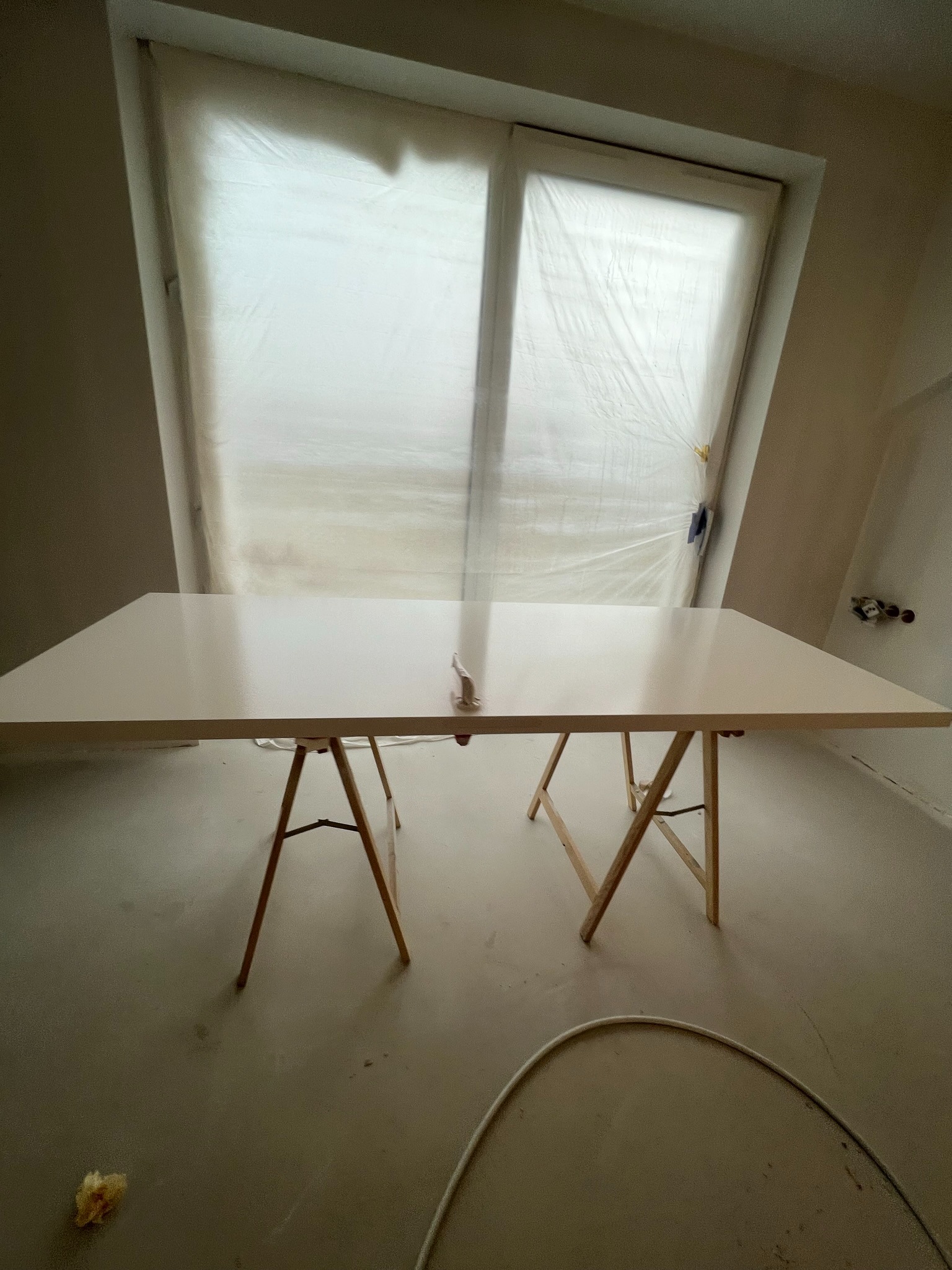 Biały blat na kozłach w pokoju z oknem zabezpieczonym folią malarską. Widoczne ślady prac wykończeniowych na podłodze. Przygotowania do malowania lub lakierowania.