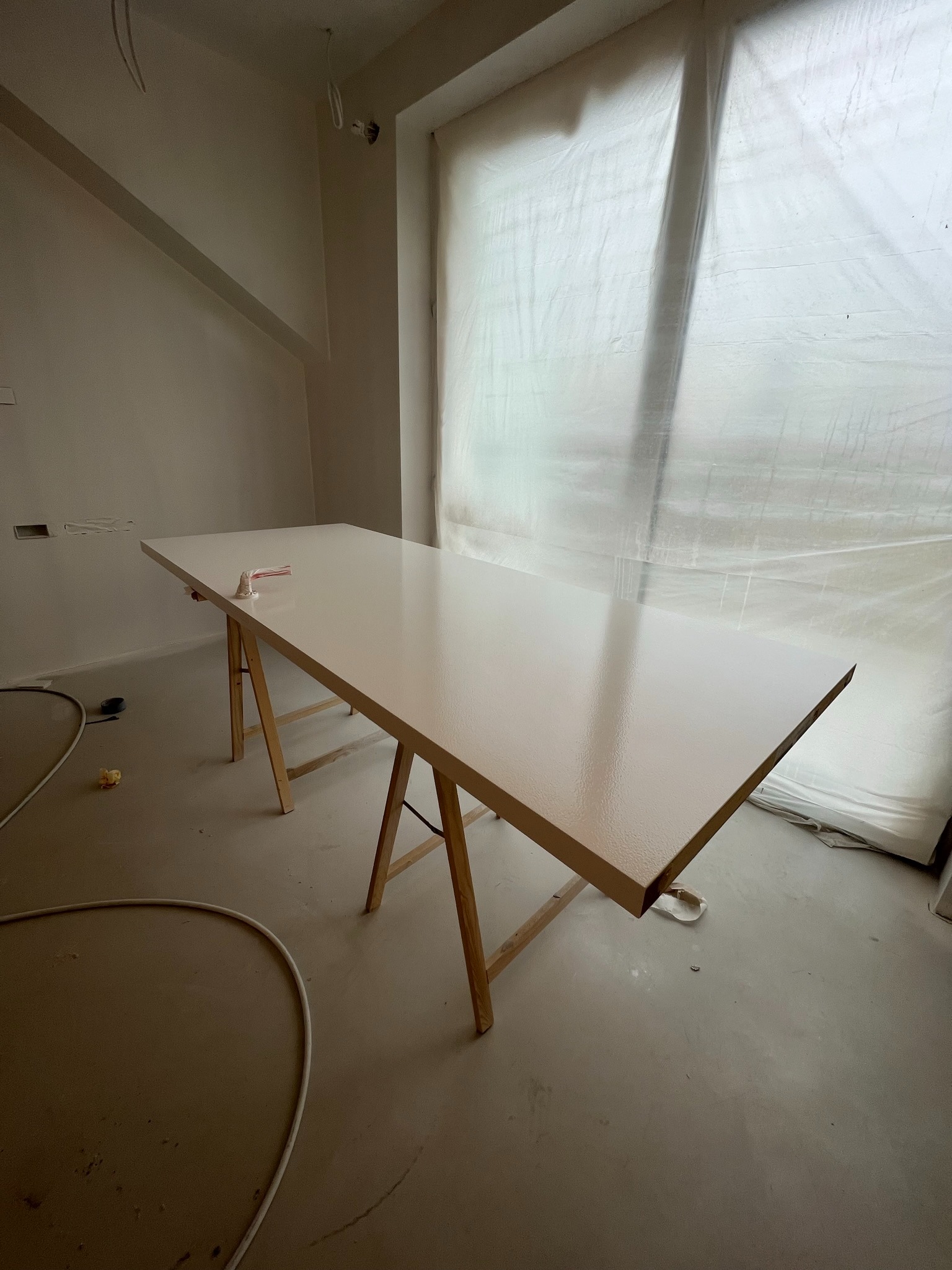 Pomalowany blat na drewnianych kozłach w pokoju zabezpieczonym folią malarską. Świeżo położona, błyszcząca farba. Widoczne narzędzia malarskie.