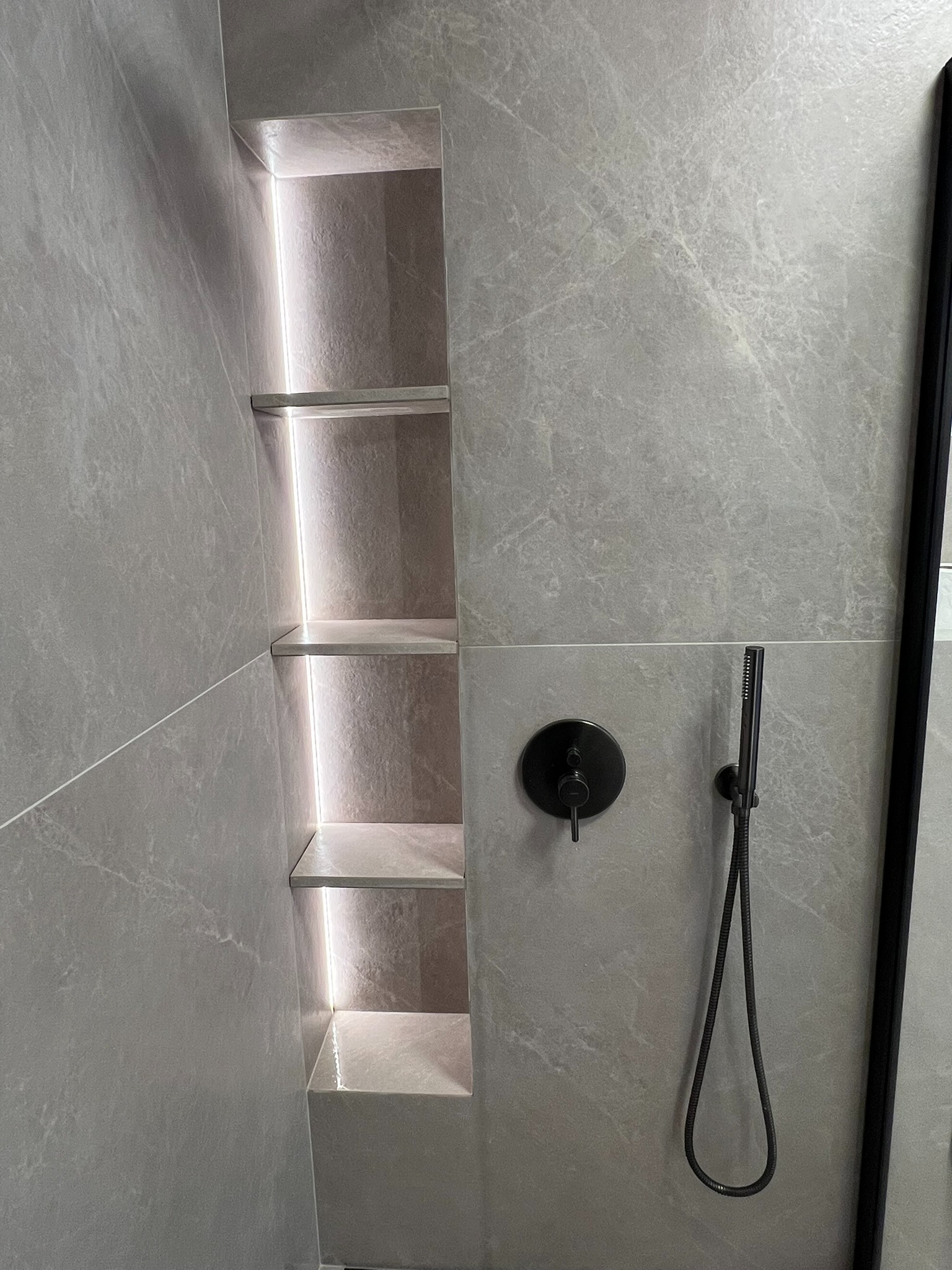 Wykończona łazienka z szarymi płytkami, wnęką z półkami podświetlonymi LED i czarną armaturą prysznicową. Minimalistyczny design, nowoczesne wykończenie.