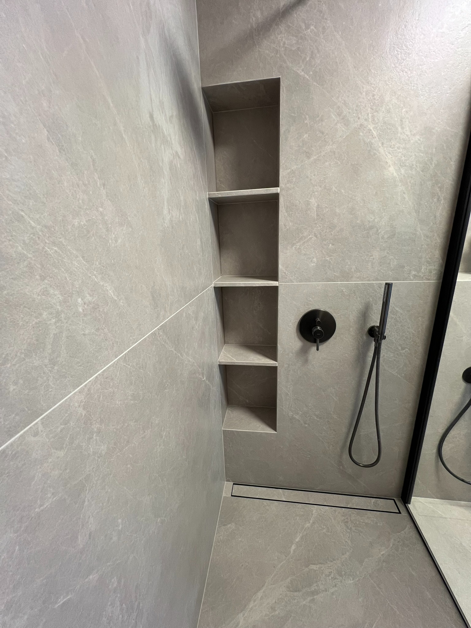 Nowoczesna łazienka z szarymi płytkami imitującymi marmur, czarną armaturą i wnęką z półkami w strefie prysznicowej, minimalistyczny design.
