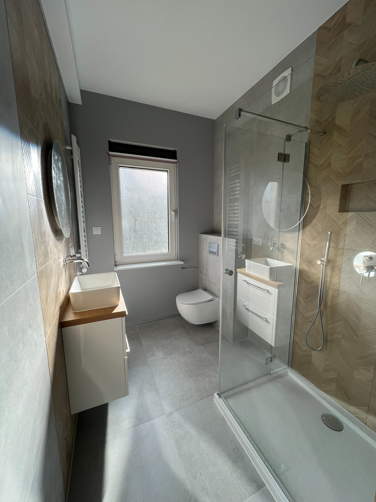 Nowoczesna łazienka z kabiną prysznicową typu walk-in, umywalką nablatową na szafce i płytkami imitującymi drewno na ścianie. Minimalistyczny design, szaro-beżowa kolorystyka.