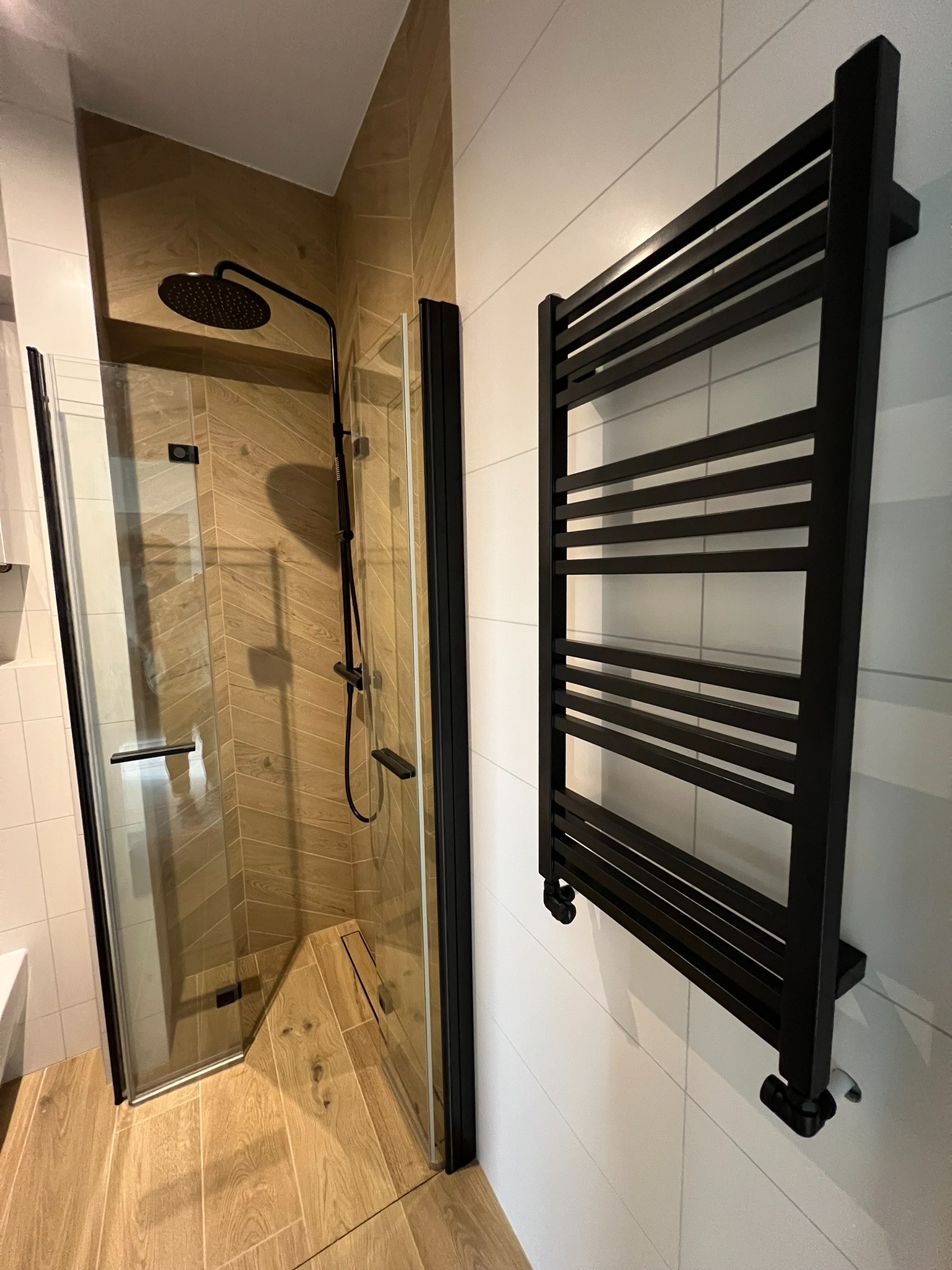 Nowoczesna łazienka z kabiną prysznicową z czarnymi ramami i grzejnikiem drabinkowym na tle białych płytek. Uwagę zwracają drewnopodobne płytki w kabinie.