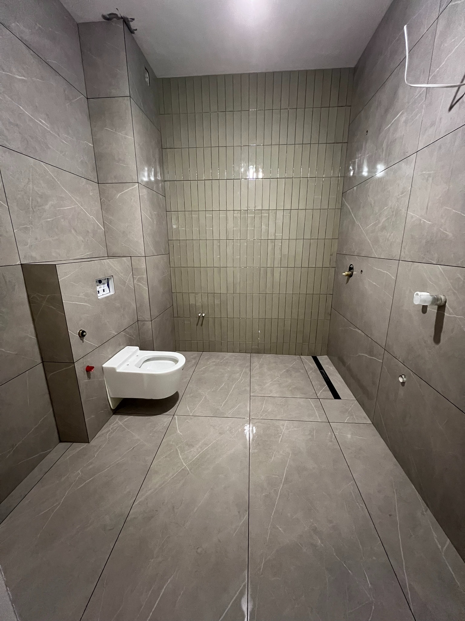 Nowoczesna łazienka w szarościach: marmurowe płytki podłogowe i ścienne, pionowe płytki na ścianie, podwieszana toaleta i minimalistyczny odpływ liniowy.