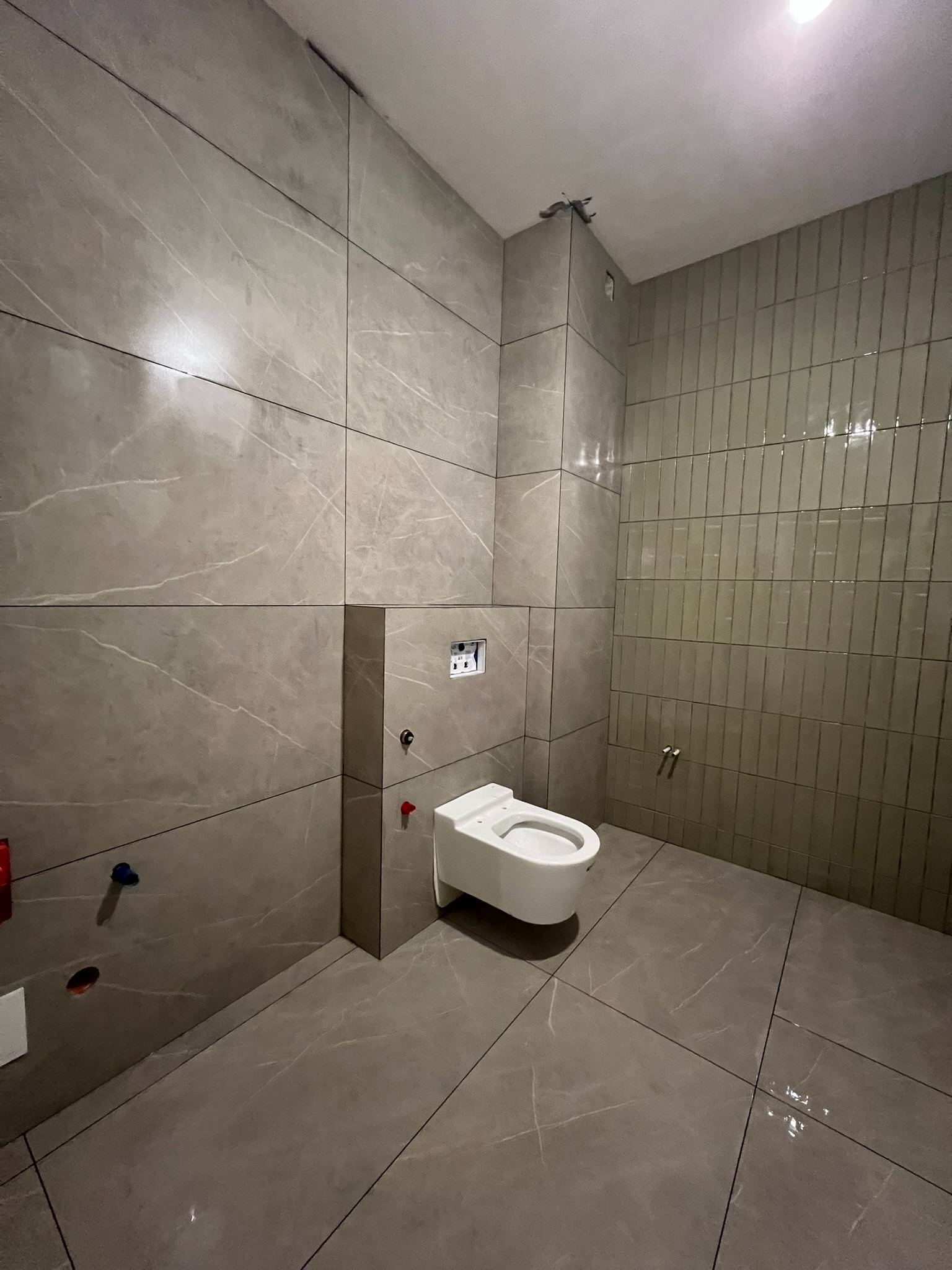 Nowoczesna łazienka z beżowymi płytkami imitującymi marmur na ścianach i podłodze. Wisząca toaleta, pionowe płytki na jednej ze ścian, sufit z punktowym oświetleniem.