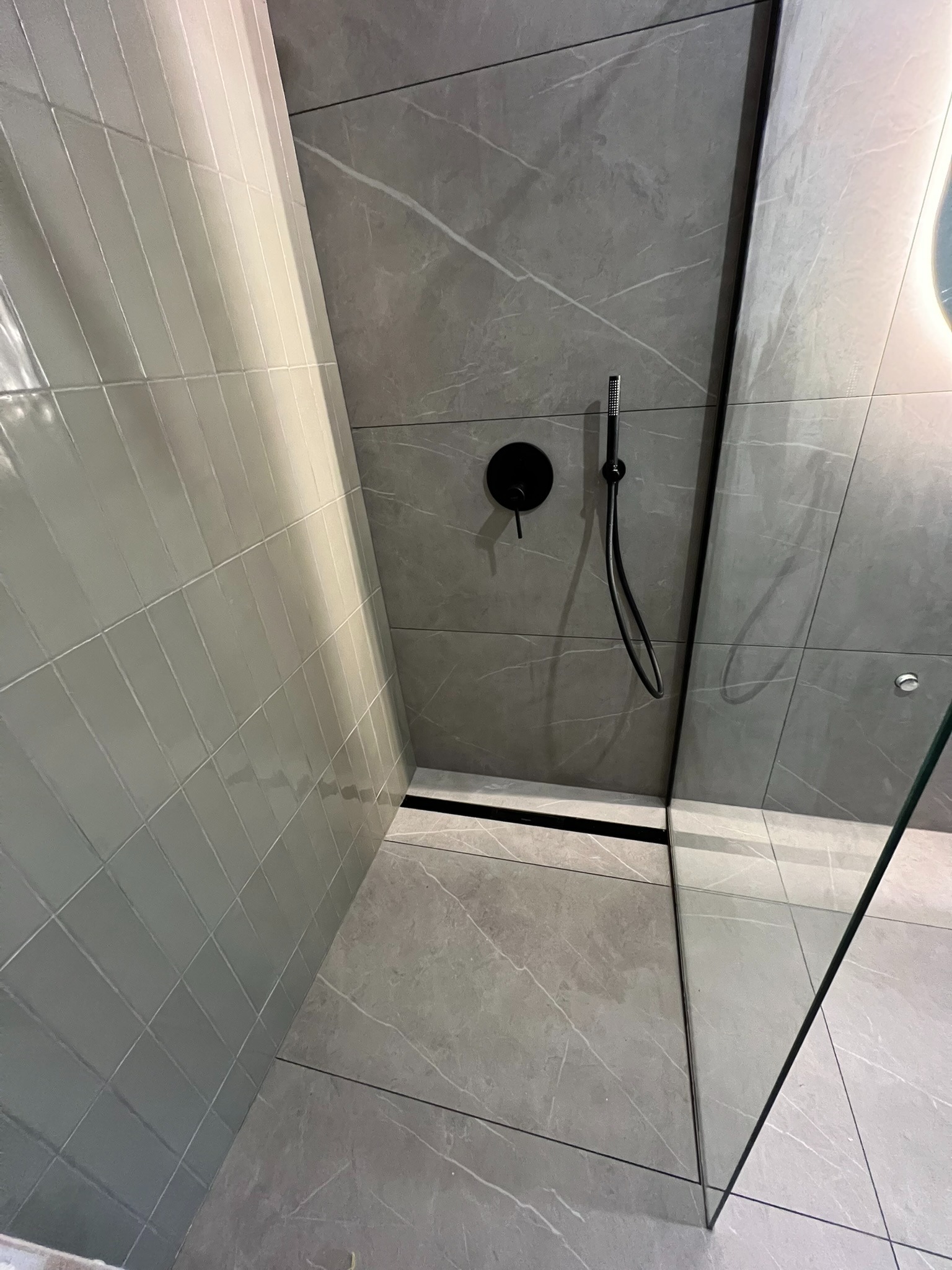 Nowoczesna kabina prysznicowa z szarymi płytkami imitującymi marmur i czarną armaturą. Minimalistyczny design z odpływem liniowym.