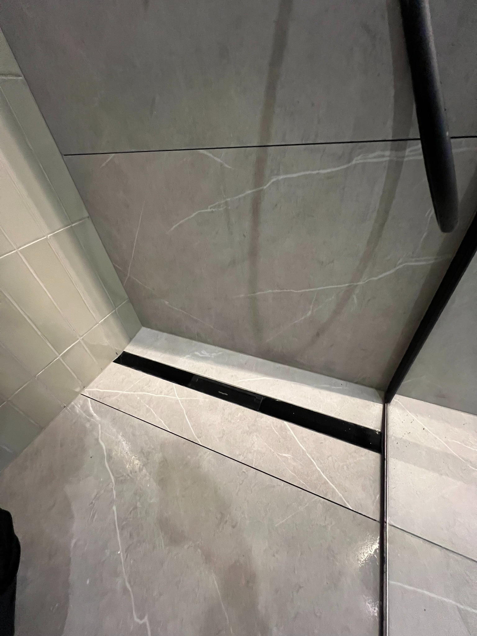 Wykończona łazienka z szarymi płytkami imitującymi marmur i czarnym odpływem liniowym. Widoczne fugi i fragment kabiny prysznicowej z czarnym wykończeniem.