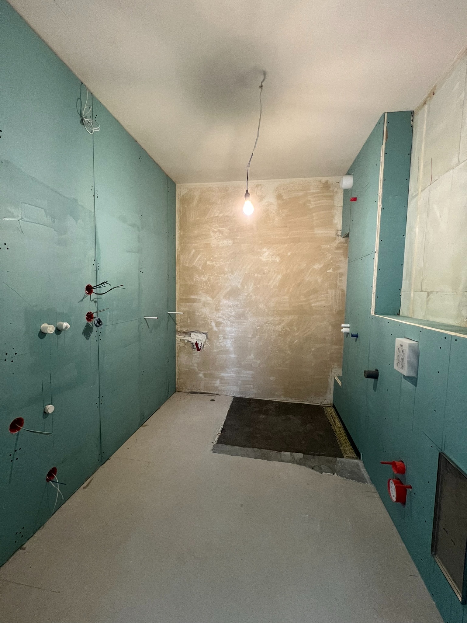 Wnętrze łazienki w trakcie remontu: ściany obłożone płytami gipsowo-kartonowymi w kolorze zielonym, widoczne instalacje wodne i elektryczne, surowa posadzka.