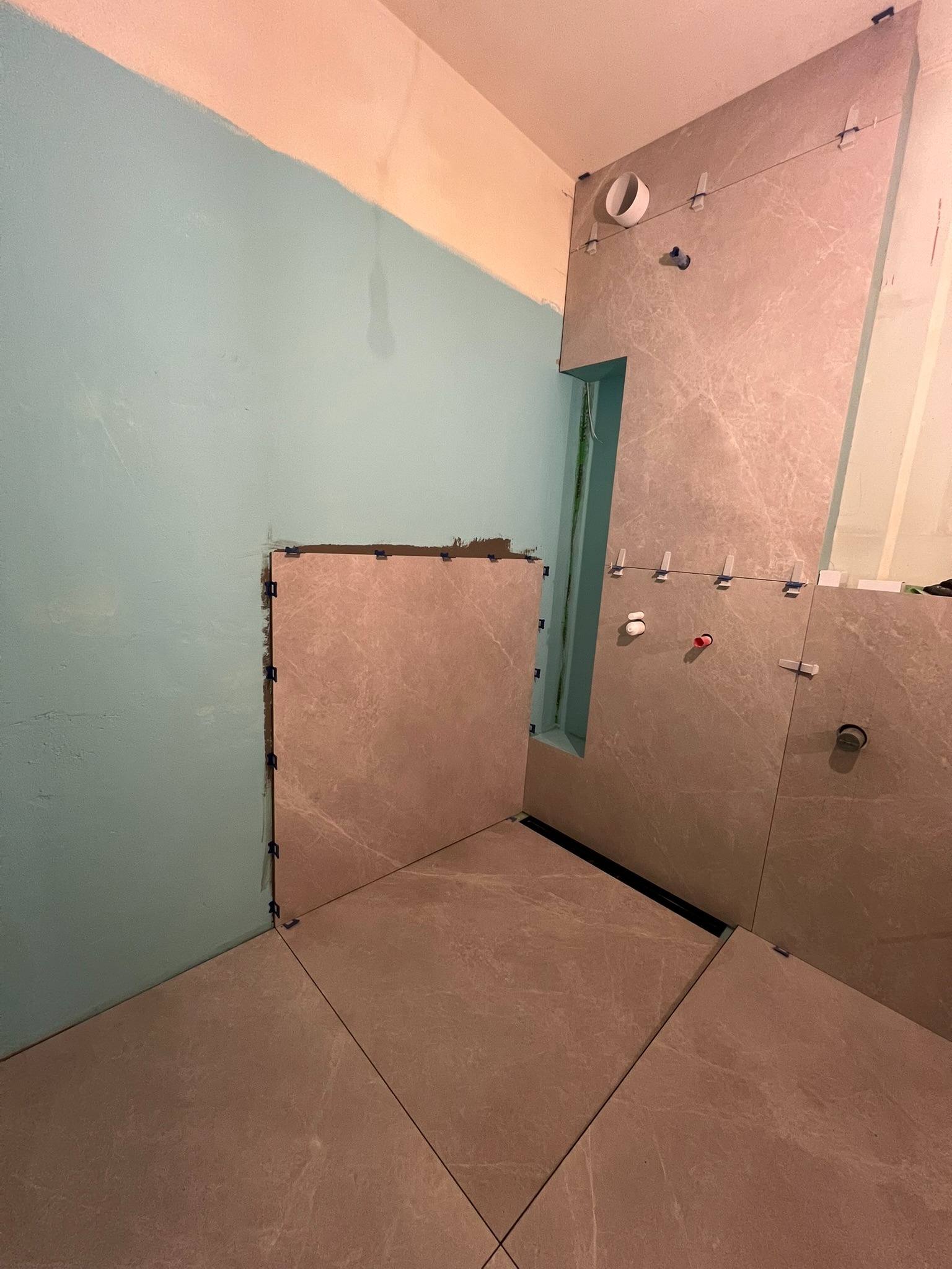 Układanie płytek w łazience: beżowe płytki imitujące marmur na ścianach, niebieska ściana częściowo wykończona, widoczny odpływ liniowy w podłodze.