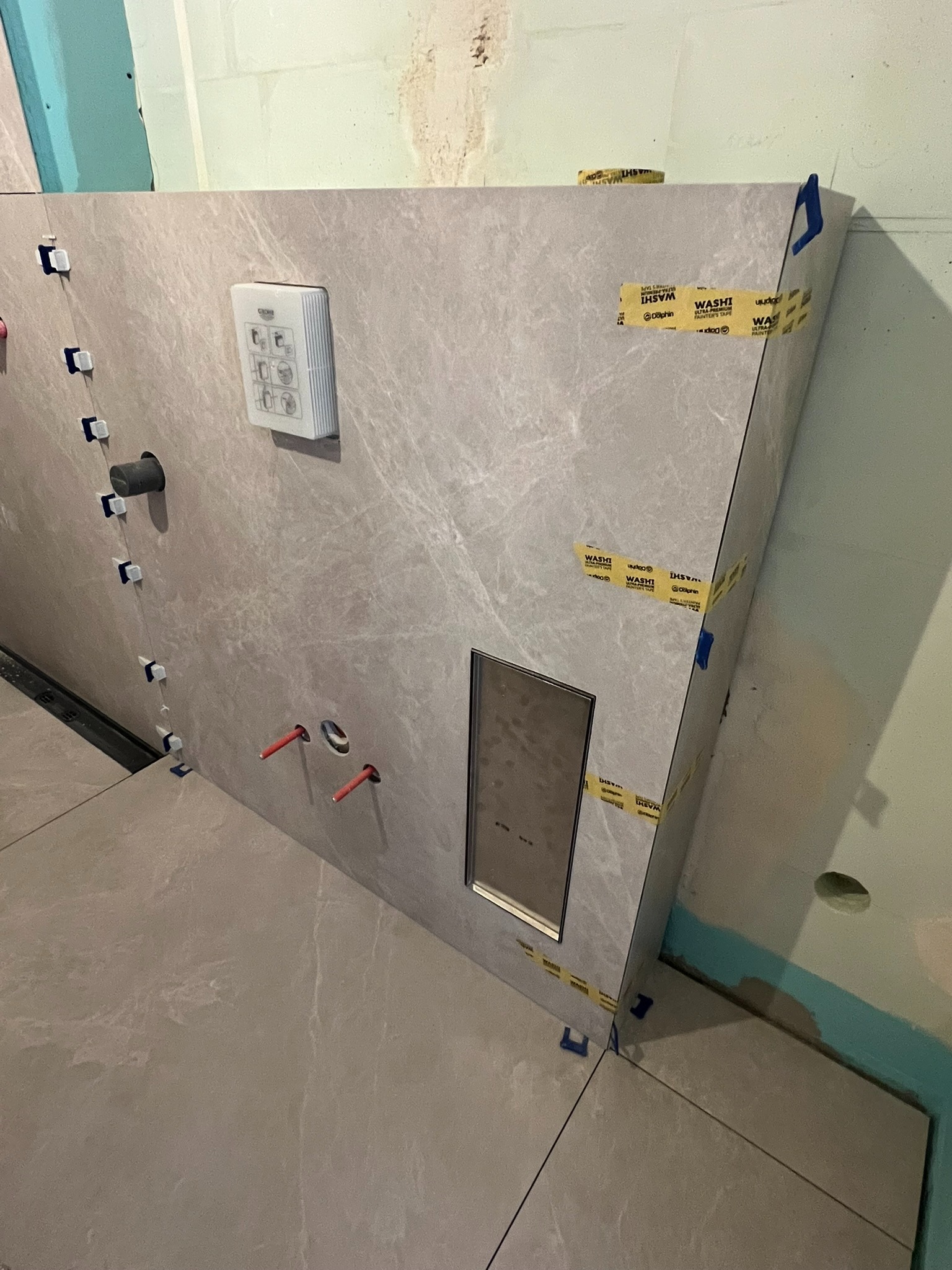 Montaż płytek imitujących marmur na ścianie z wnęką i armaturą w łazience. Widoczne poziomowanie i taśmy malarskie. Detal wykończenia.