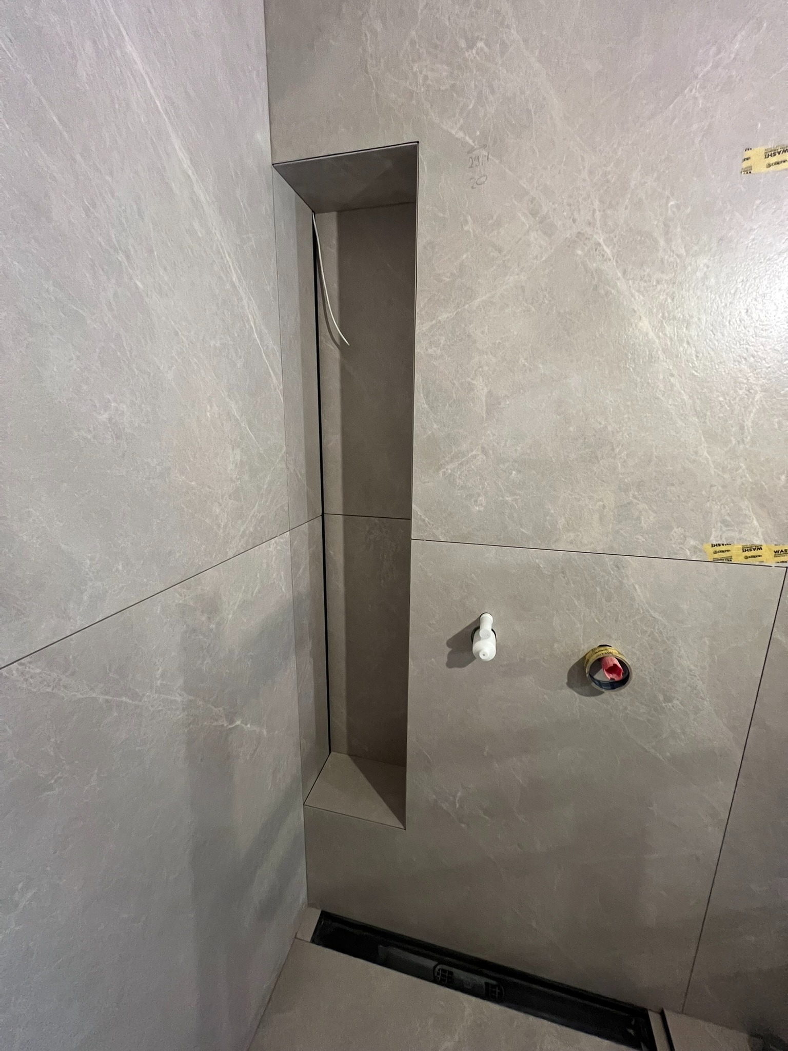 Wykończona wnęka prysznicowa z szarymi płytkami imitującymi marmur, widoczne elementy instalacji wodnej i odpływ liniowy w podłodze. Minimalistyczny design łazienki.