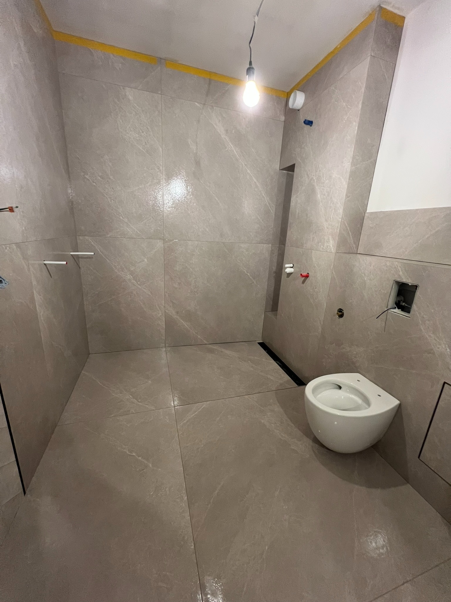 Wykończona łazienka z szarymi płytkami imitującymi marmur, wisząca toaleta, odpływ liniowy. Żółta taśma malarska wzdłuż sufitu.