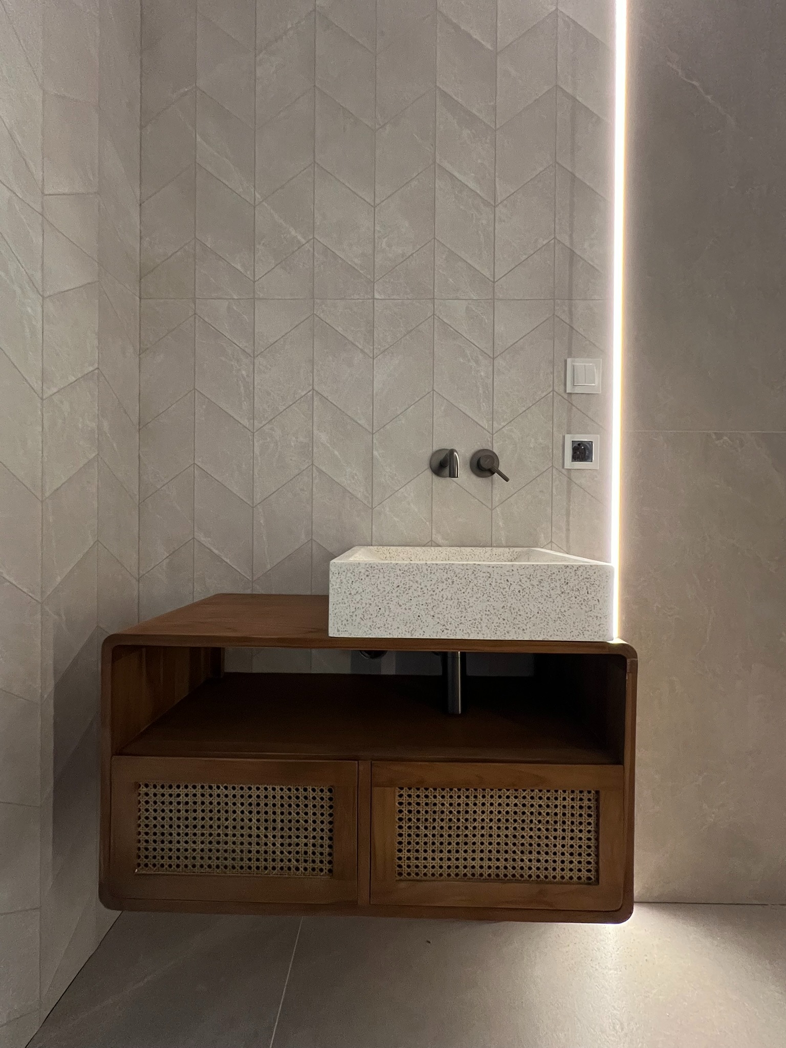 Nowoczesna łazienka z umywalką z lastryko na drewnianej szafce z rattanowymi frontami. Ściana wyłożona płytkami w jodełkę i pionowe oświetlenie LED.