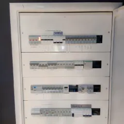 Otwarta, biała skrzynka rozdzielcza z wyłącznikami nadprądowymi i opisami obwodów, w tym 'Światło', 'Kuchnia', 'Wentylator', 'Dźwięk', z jednym wyłącznikiem w pozycji włączonej, wskazującym...